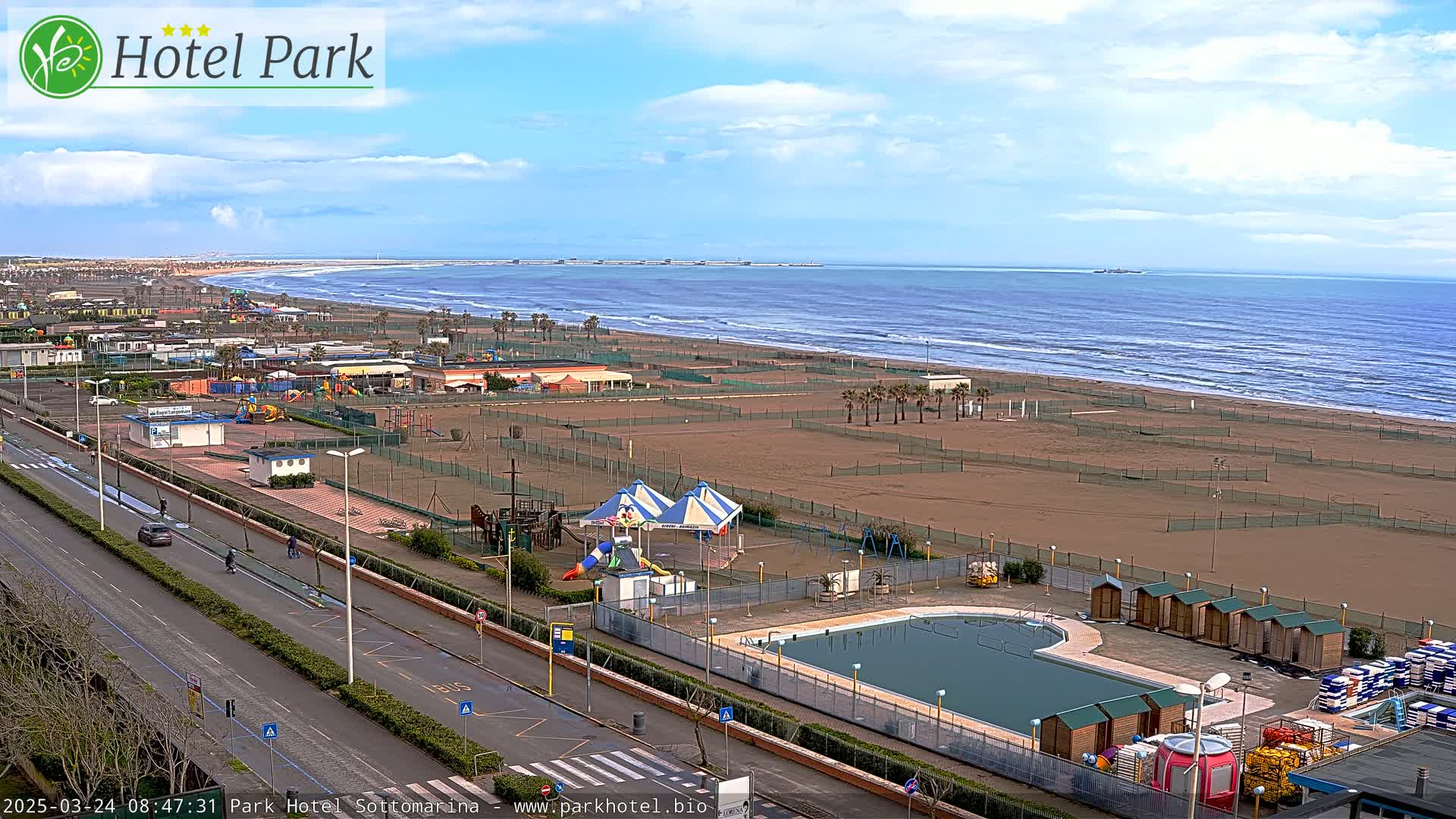 An aerial view shows a sandy beach with a calm ocean under a partly cloudy sky, adjacent to a beachfront area with a swimming pool, playground, and small buildings.