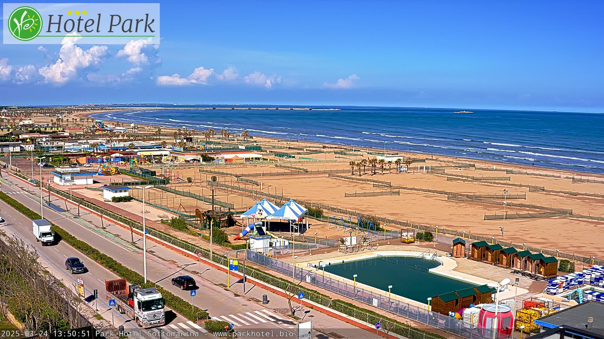 An aerial view shows a beach with a playground, swimming pool, and a road with vehicles, under a mostly sunny sky with some clouds.