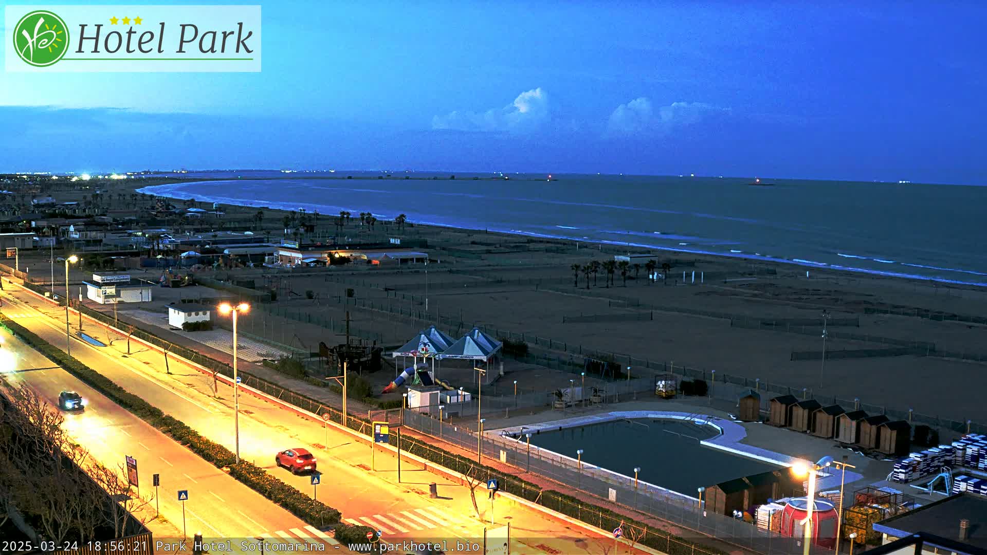 A nighttime view of a beach with a calm sea under a twilight sky, showing a road, buildings, a swimming pool, and a playground.