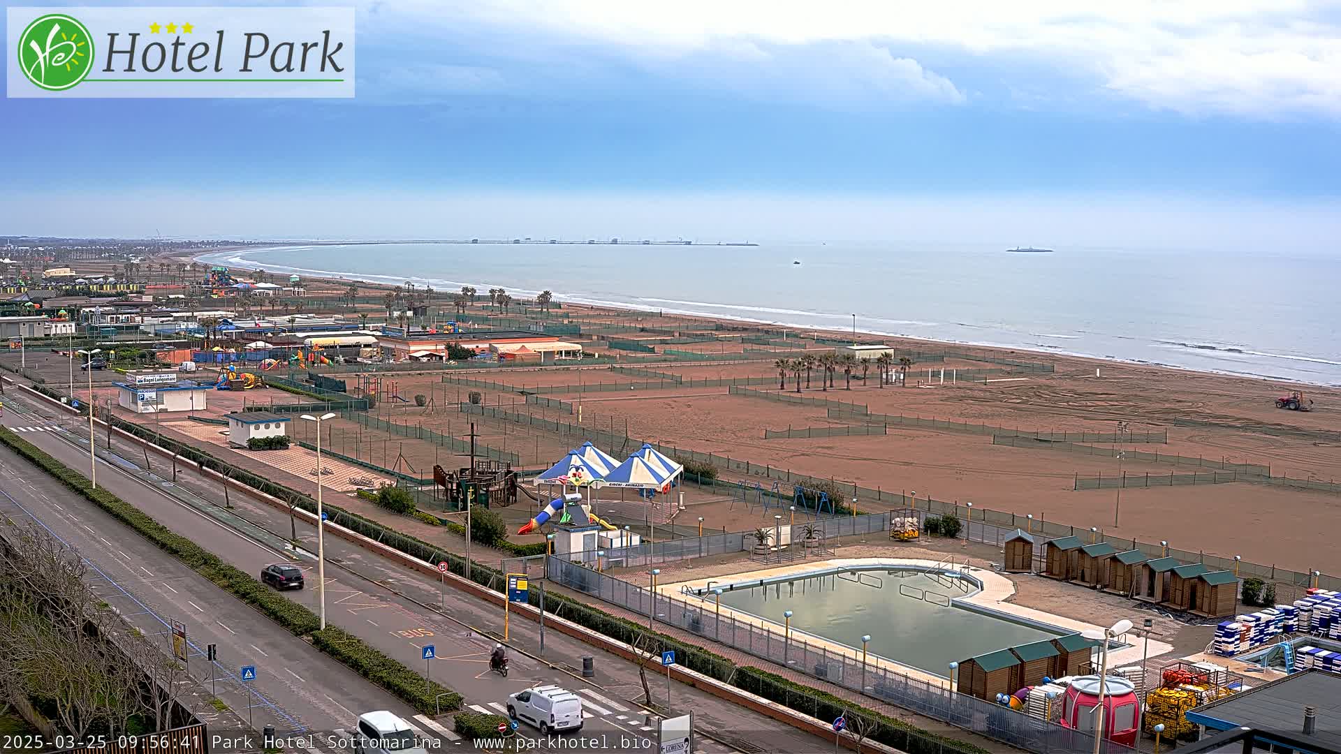 An aerial view of a beach with a swimming pool, playground equipment, and a road, under a partly cloudy sky.
