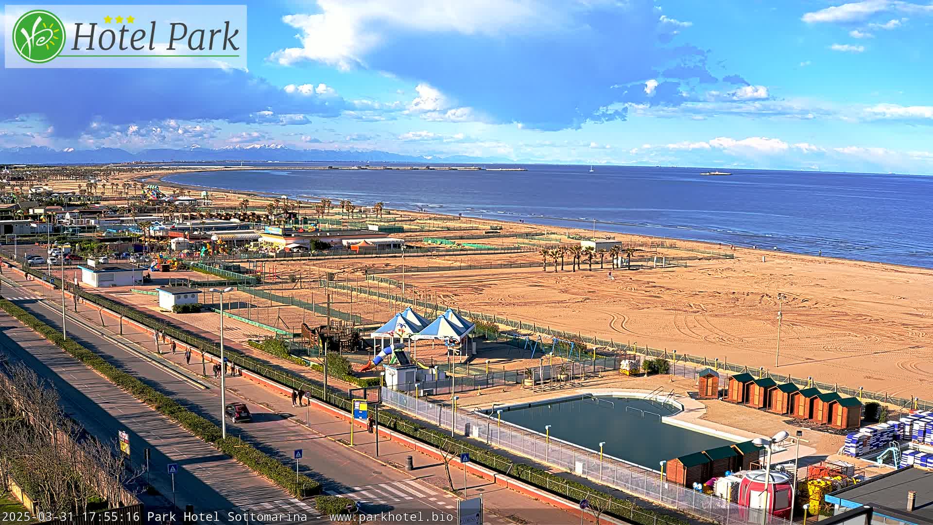 A beach with a swimming pool, playground equipment, and beach huts is visible under a partly cloudy sky, with mountains in the distance.