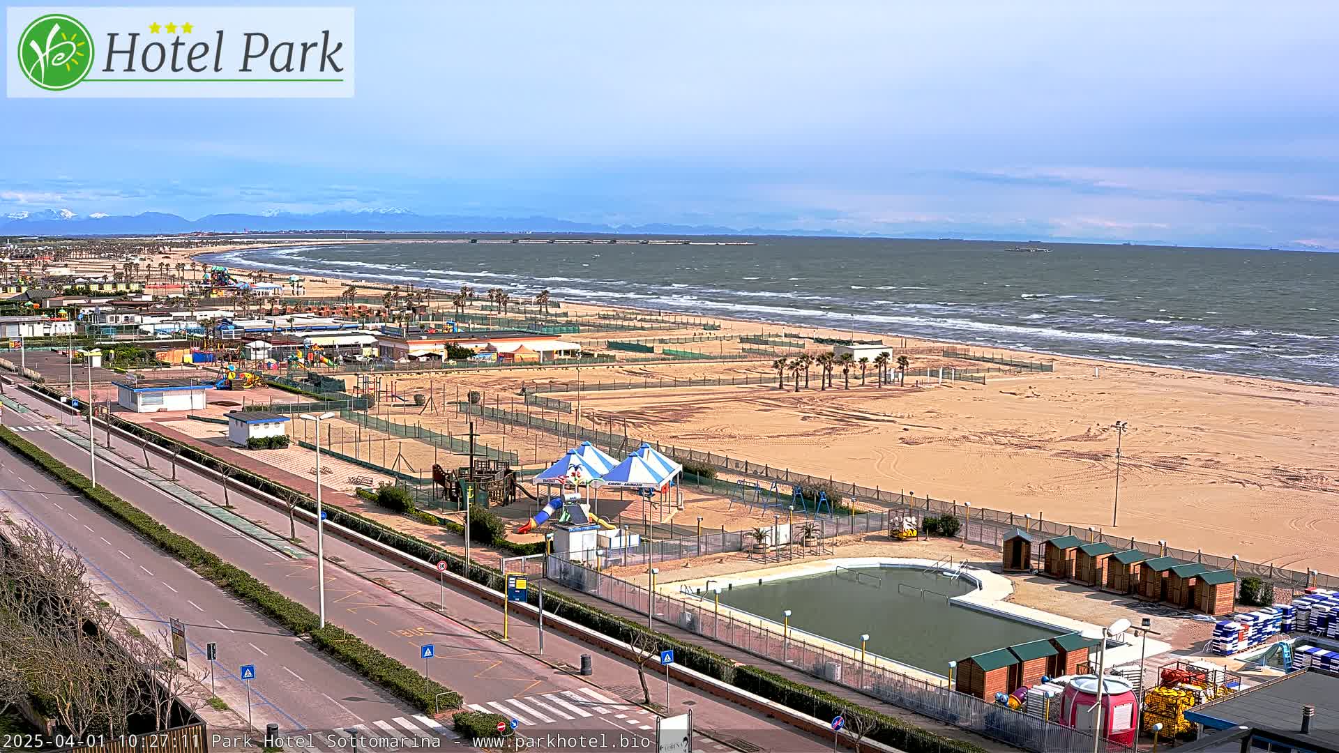 An aerial view shows a sandy beach with a playground, pool, and small buildings under a partly cloudy sky with a windy ocean in the background.