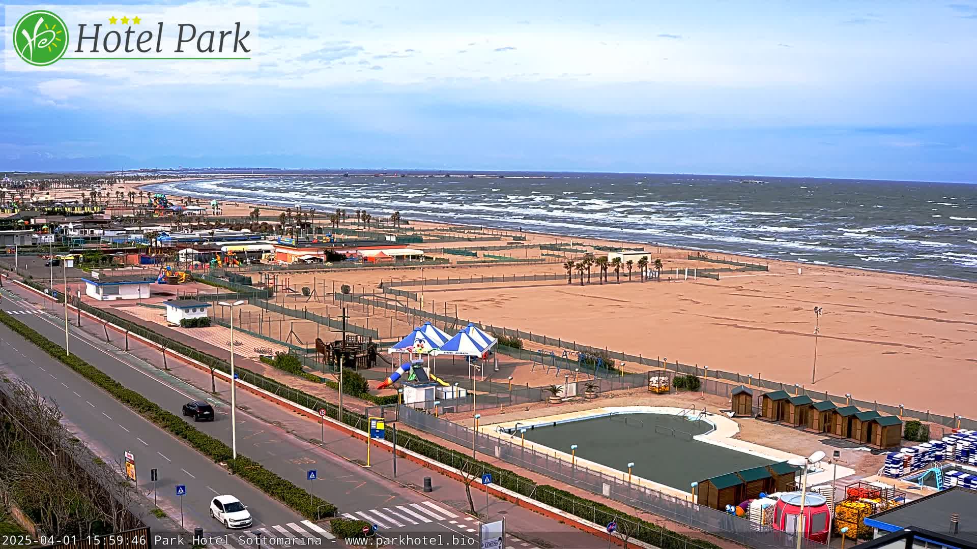 An aerial view shows a beach with choppy waves, a road with cars, and a beachfront area with a pool, playground equipment, and beach huts.