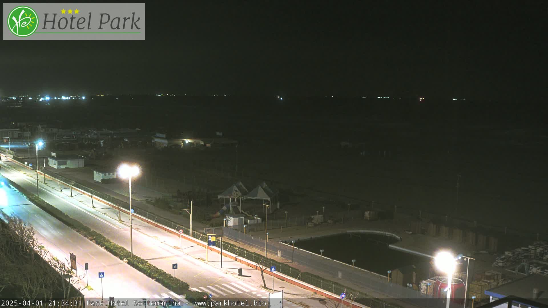 A nighttime, long shot of a mostly empty beach and roadway with some lights and a small playground visible.