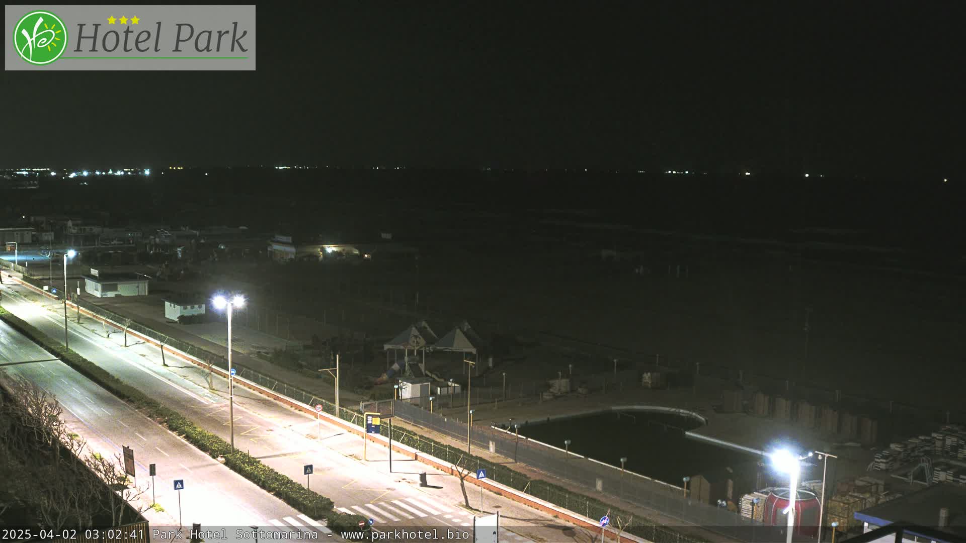 A nighttime view of a coastal road, beach, and playground area, with streetlights illuminating the scene.