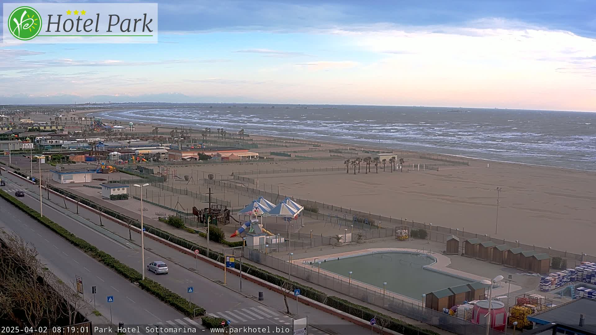 A seaside resort area with a beach showing choppy waves, a playground, a pool, and a road, under a partly cloudy sky.