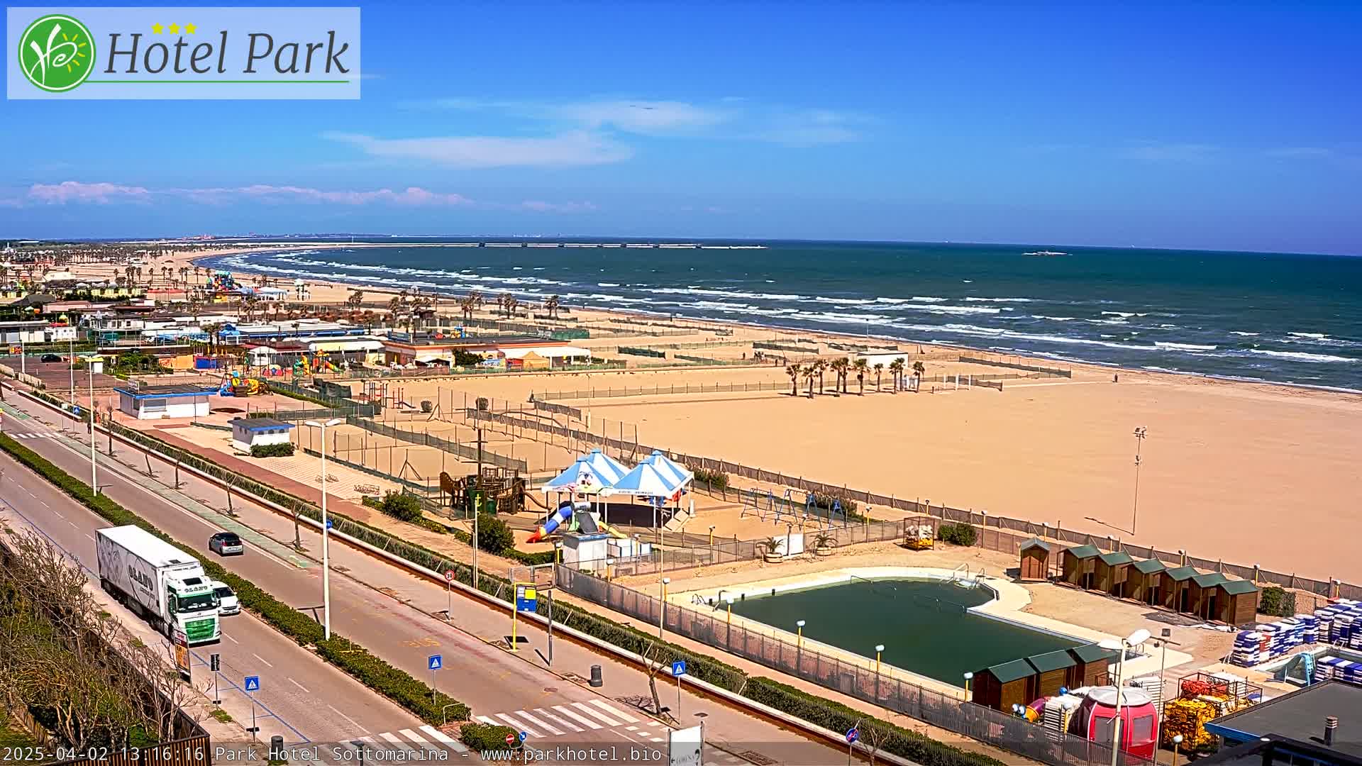 A sandy beach with a playground and a small pool is next to a road with vehicles, all under a sunny sky with some clouds and a wavy ocean in the background.