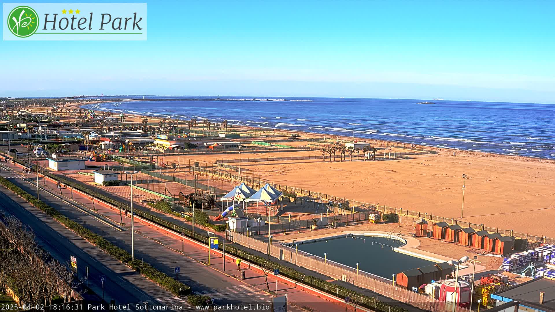 A sandy beach with a calm ocean, a beachfront road with a swimming pool and playground next to it, under a clear sunny sky.