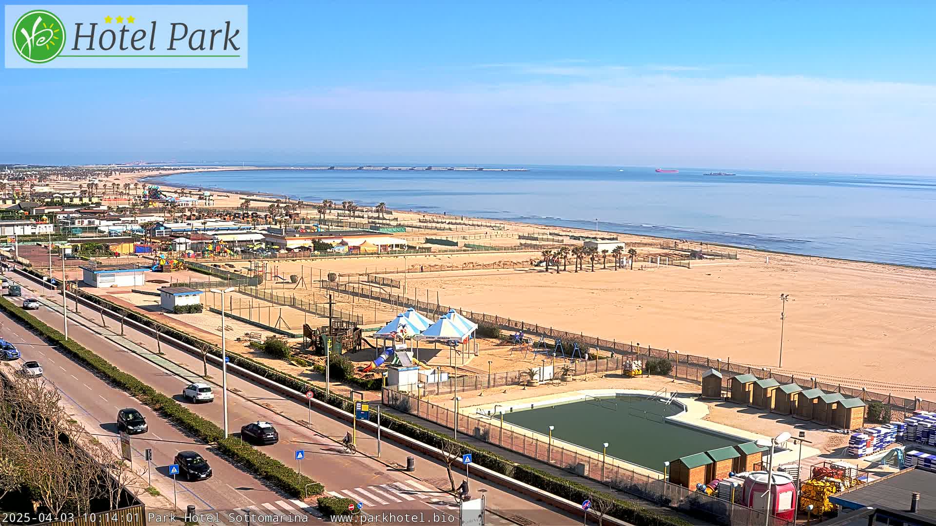 A sandy beach stretches along a calm ocean under a clear blue sky, with a beachfront road featuring cars and various playground equipment and a pool visible nearby.