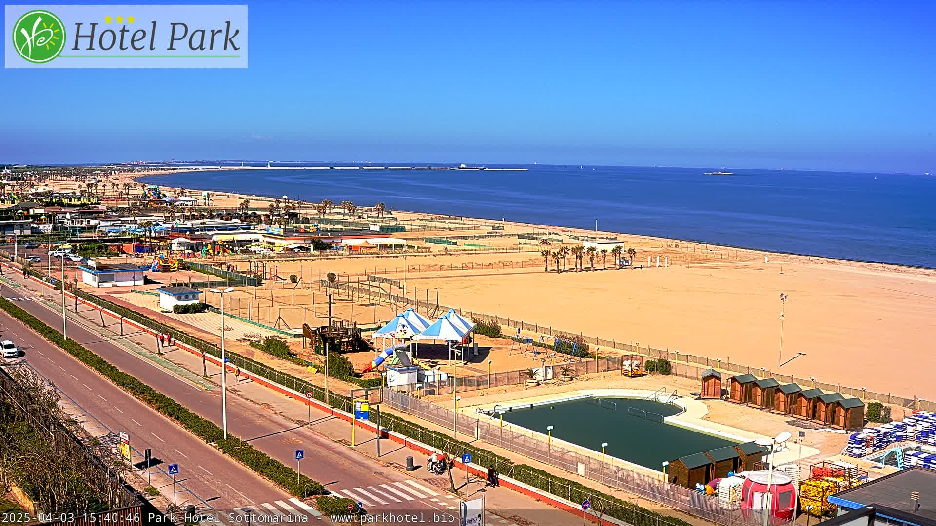 A sandy beach with a playground, swimming pool, and small buildings stretches along a calm blue ocean under a clear blue sky.