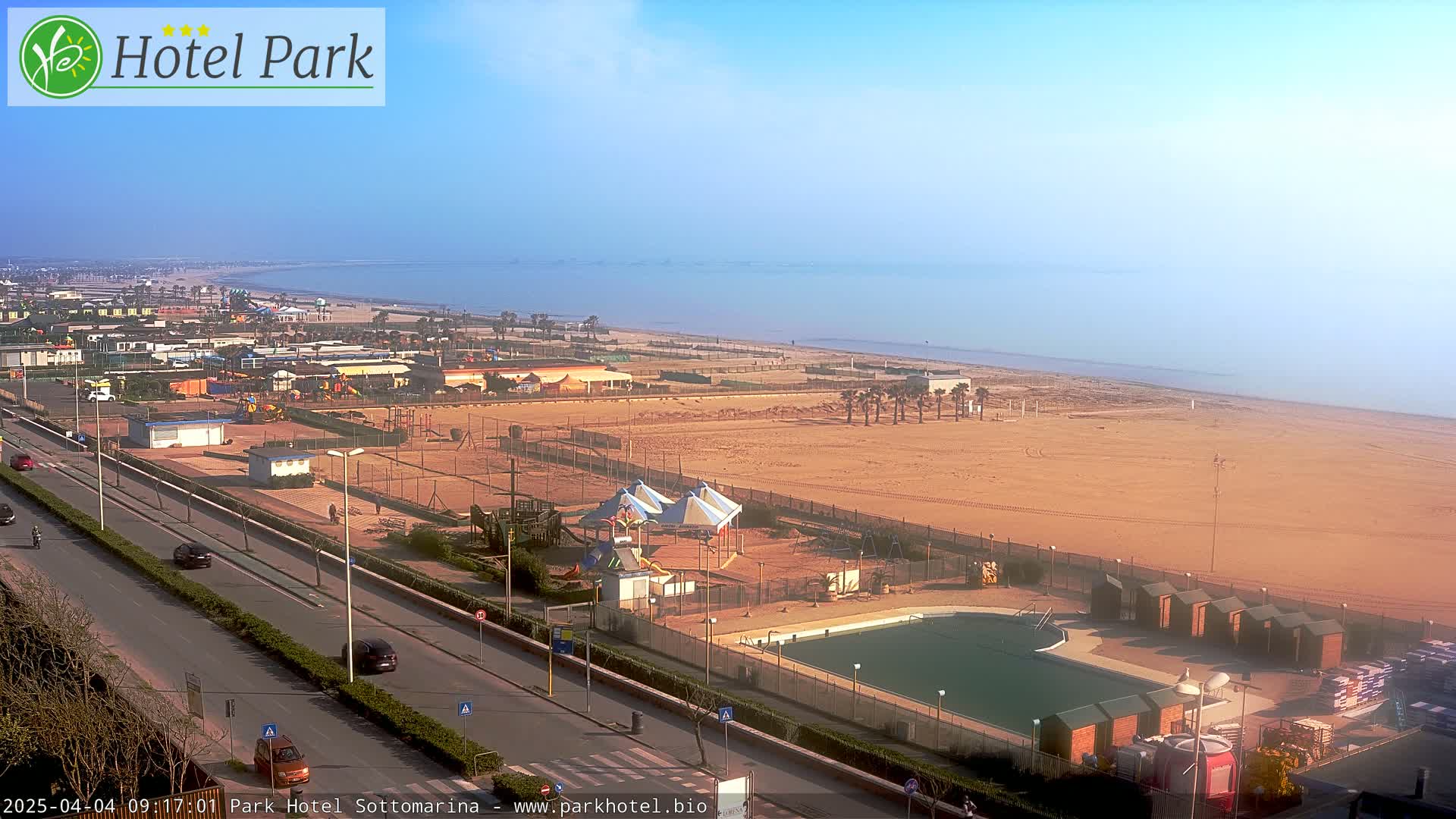 A sandy beach with a swimming pool, playground, and several buildings is visible from an elevated viewpoint on a clear, sunny day.