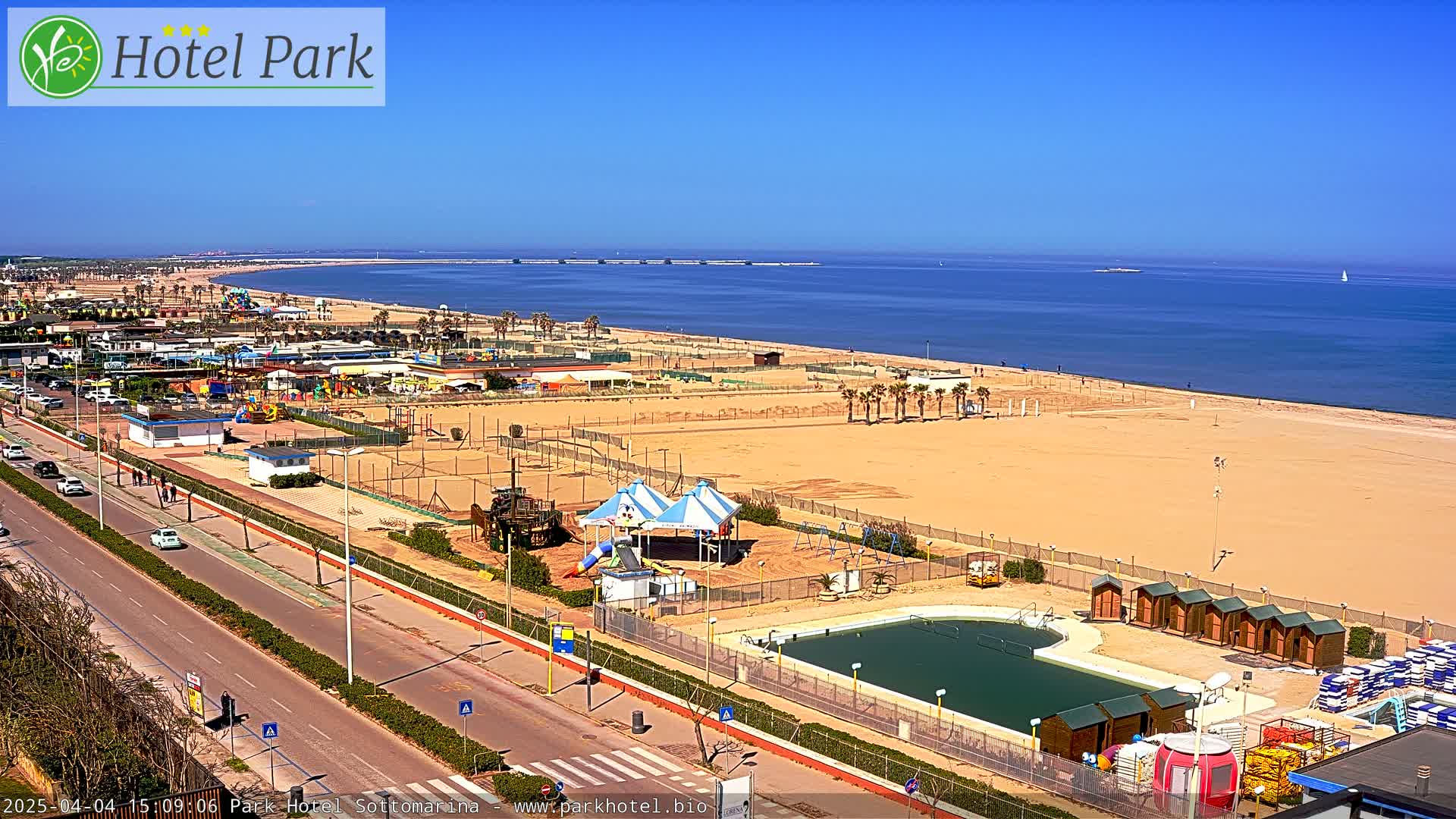 A sandy beach with a playground, swimming pool, and a road running alongside it on a sunny day.