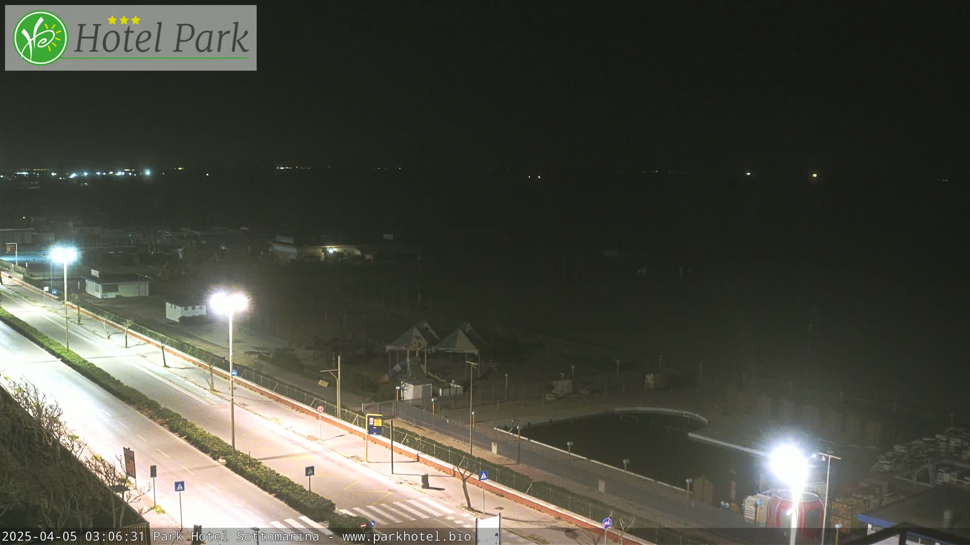 A nighttime view of a coastal road with streetlights illuminating a mostly empty beach area and a few buildings.