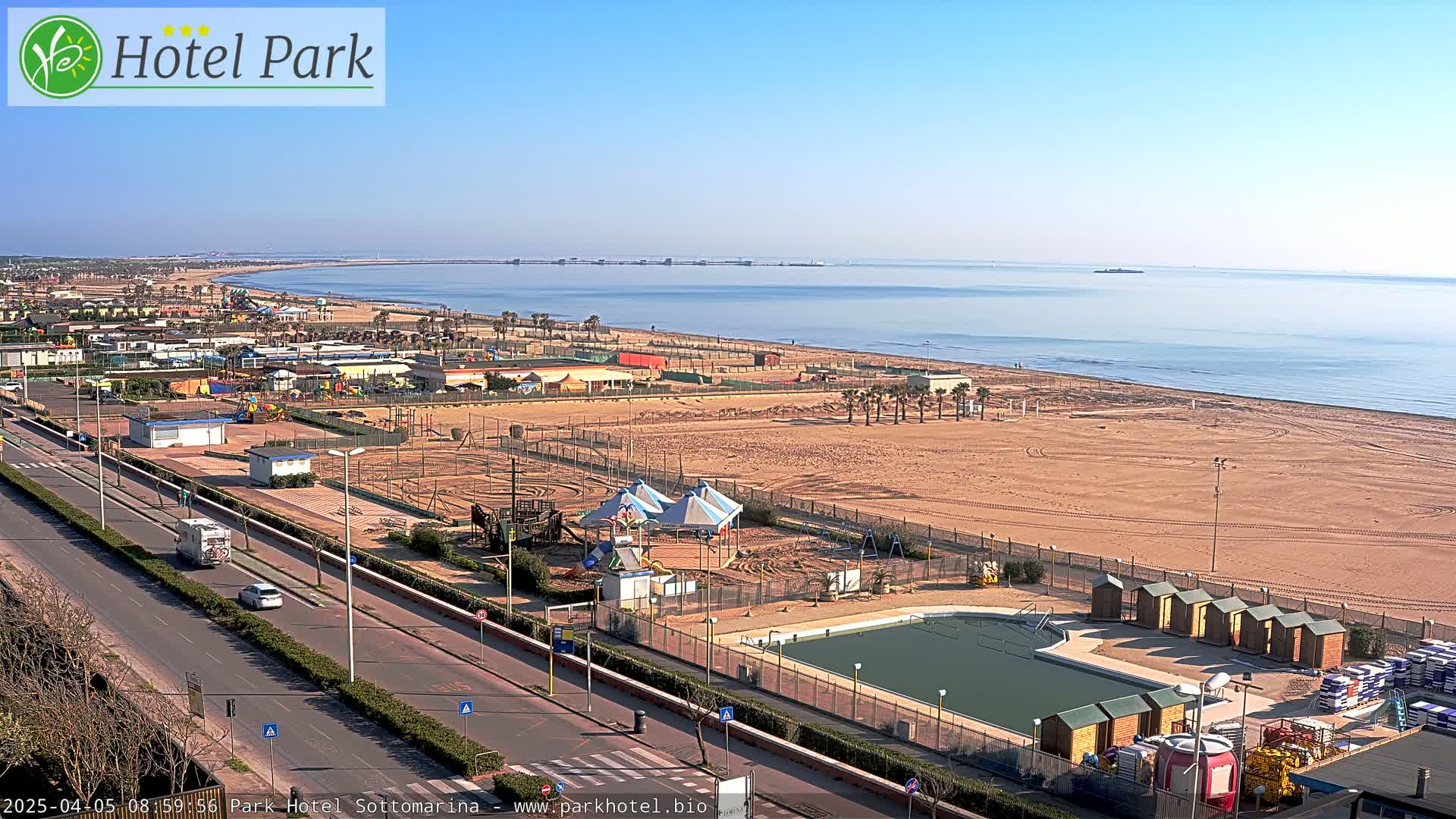 A wide shot of a beachside area on a sunny day shows a sandy beach, a road with cars, a swimming pool, and various playground equipment.