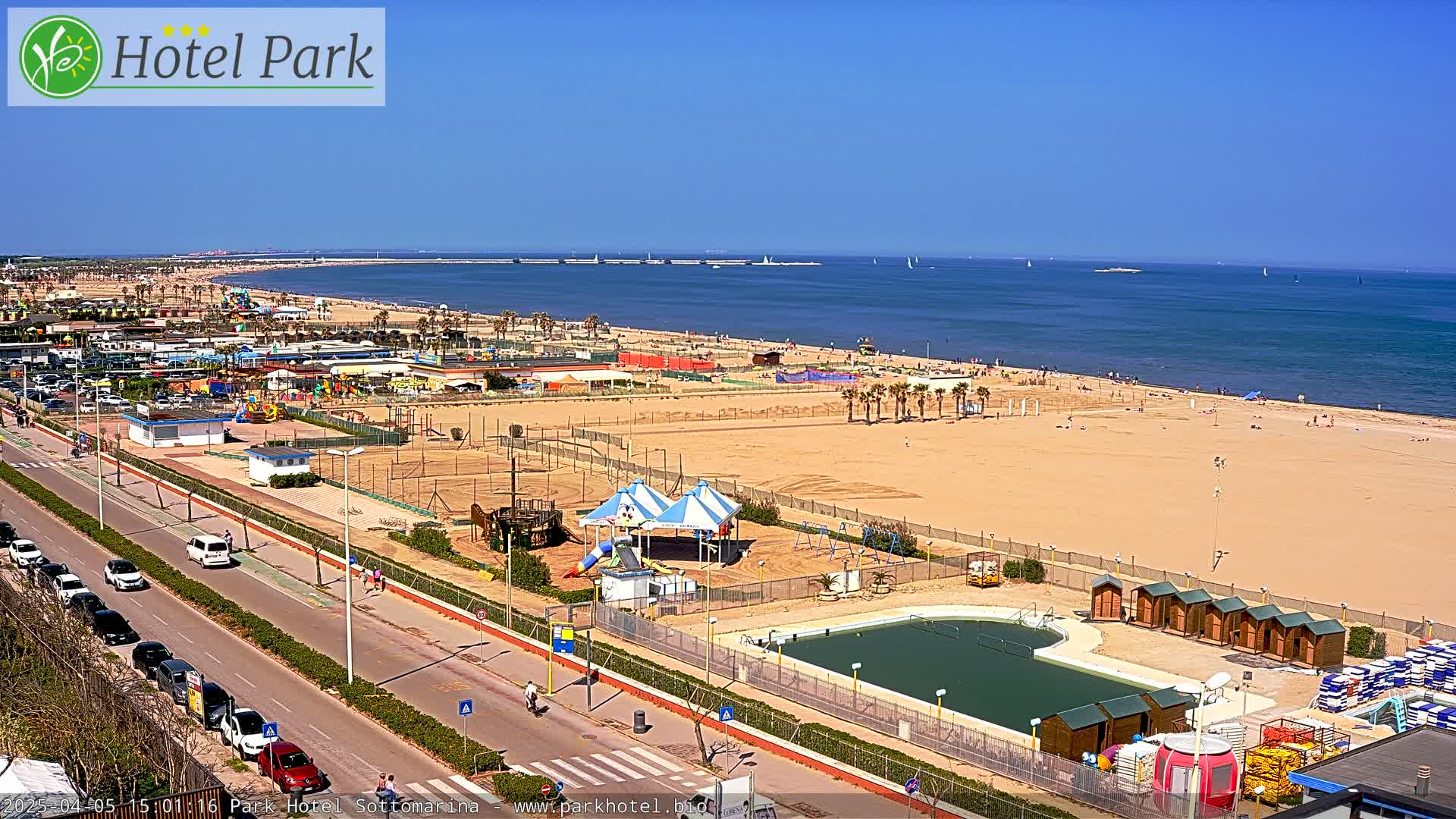 An aerial view of a beach on a sunny day shows a sandy shore, a swimming pool, playground equipment, and a road with cars parked along the side.
