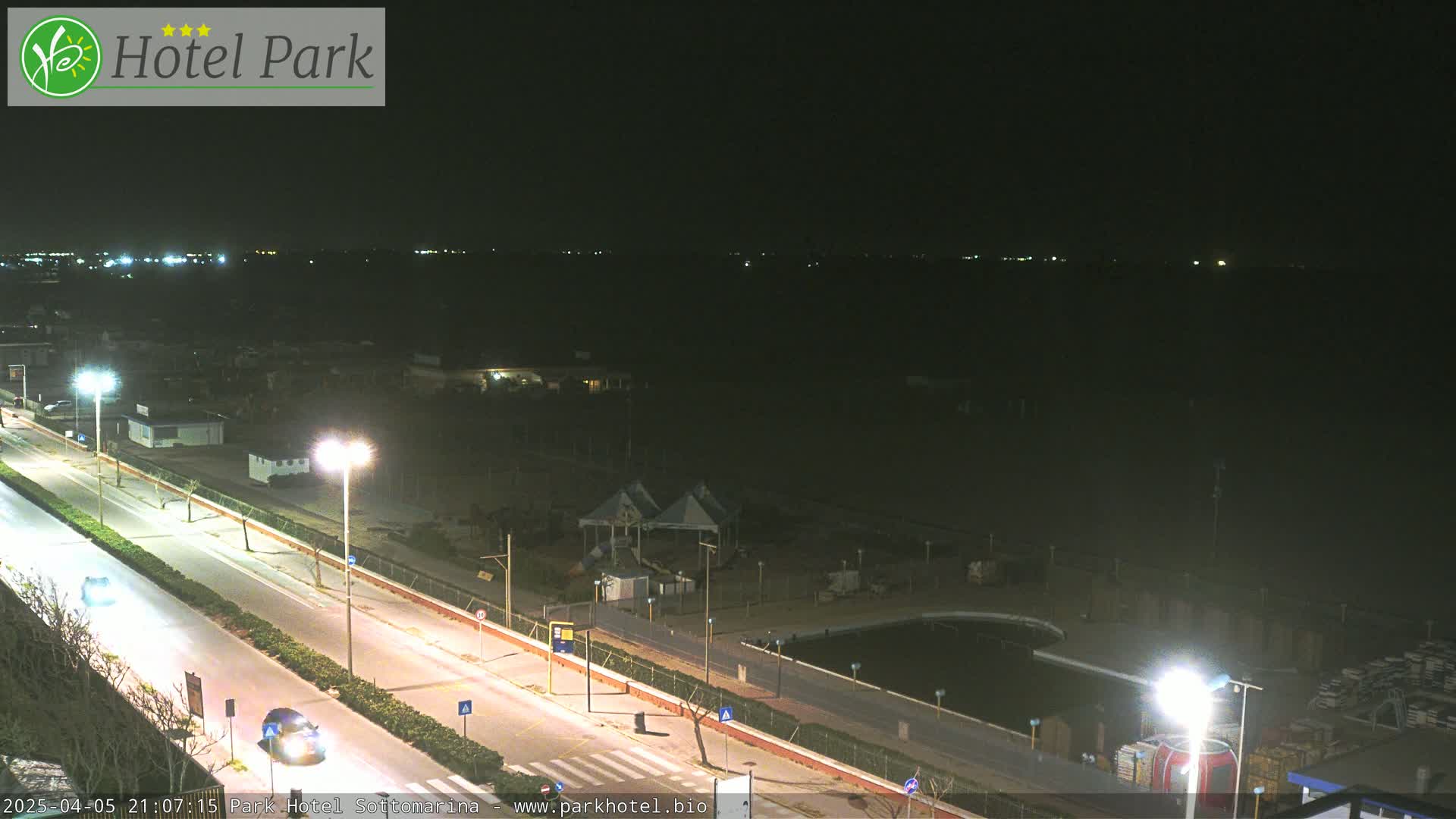 A nighttime view of a coastal road with beachfront buildings and a dark, calm body of water in the background.
