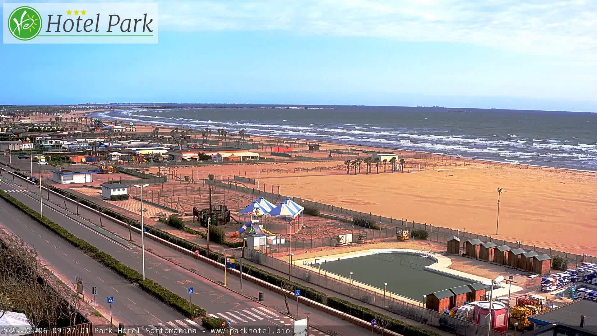 A sandy beach with a playground, swimming pool, and row of changing rooms is visible next to a road, all overlooking a windy ocean.
