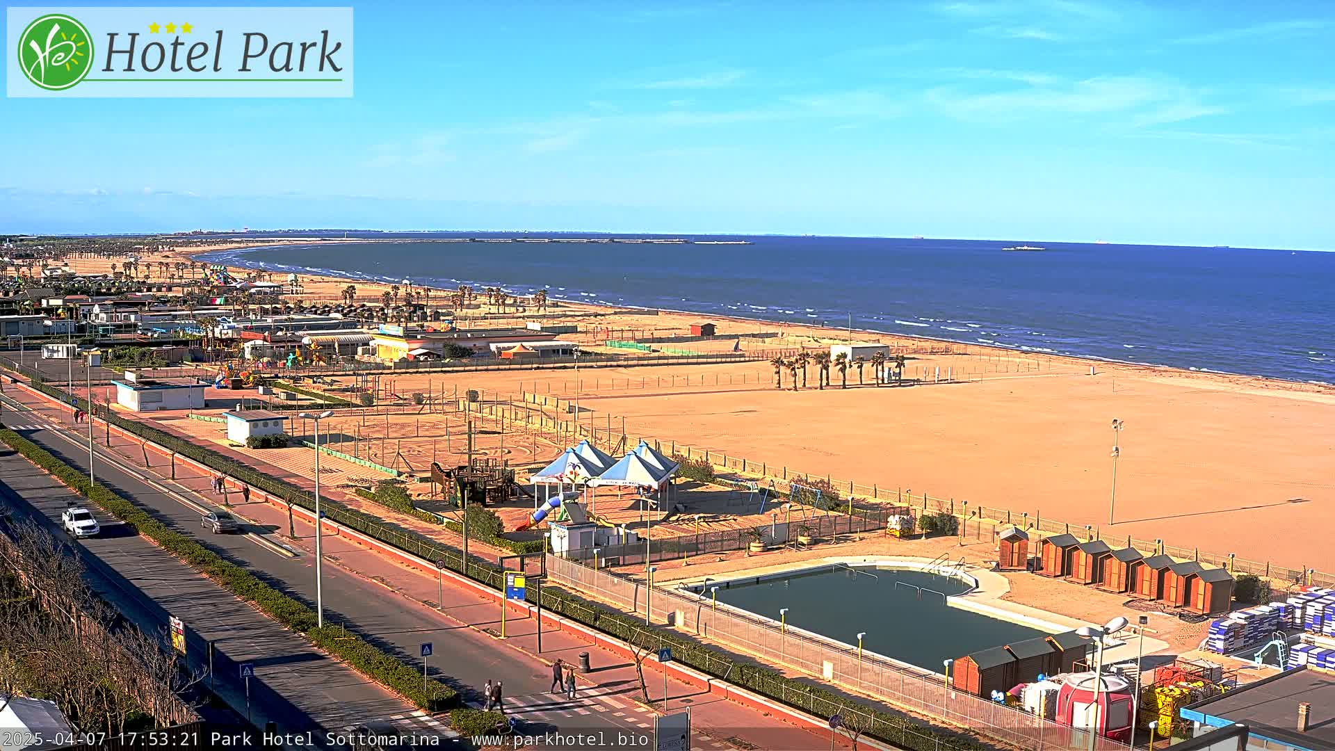A beach with a playground, swimming pool, and small buildings is visible, next to a road with cars and pedestrians, all under a clear, sunny sky.