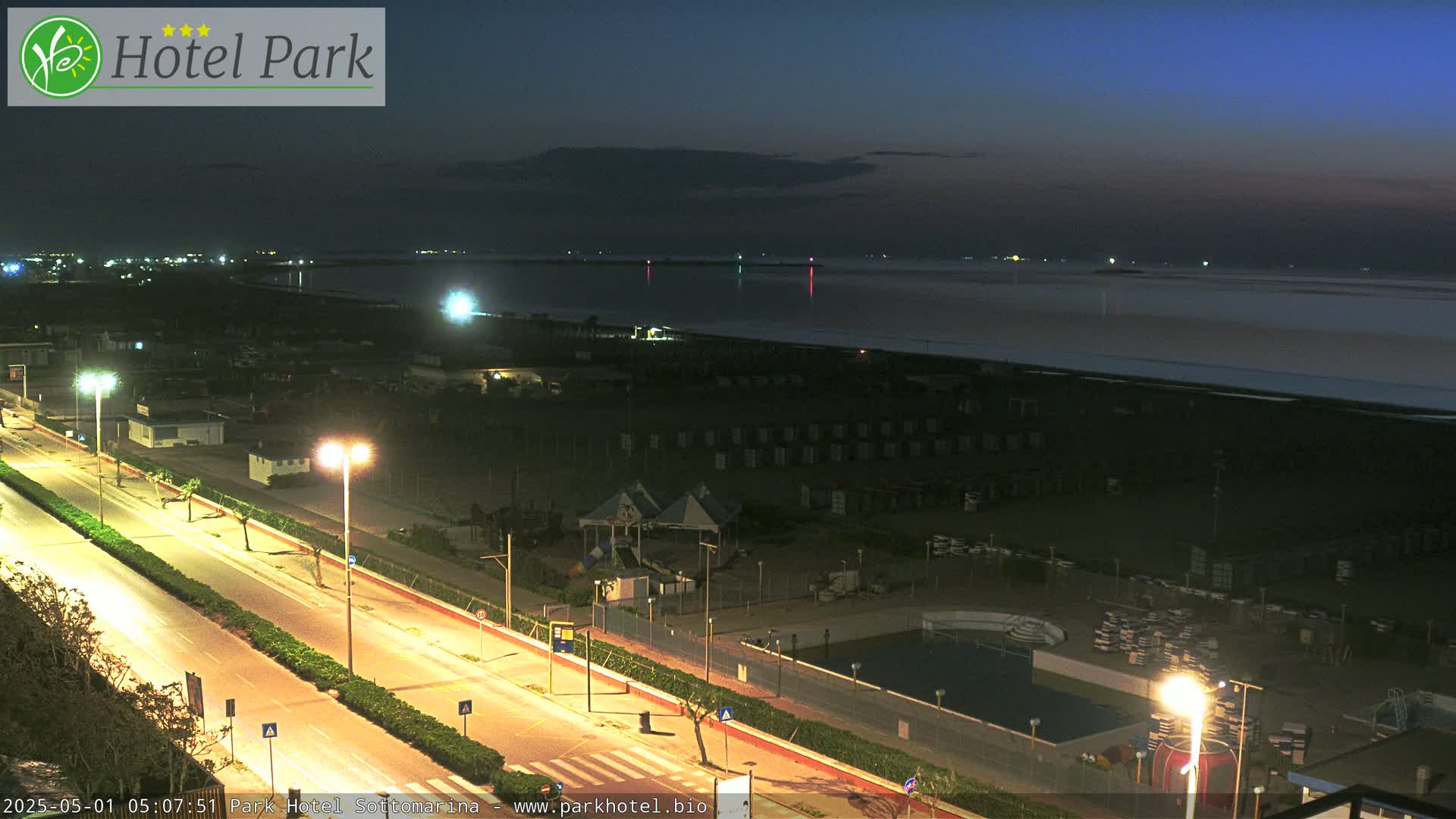 A nighttime view of a beach with rows of beach huts, a road with streetlights, and a swimming pool, under a dark blue, twilight sky.