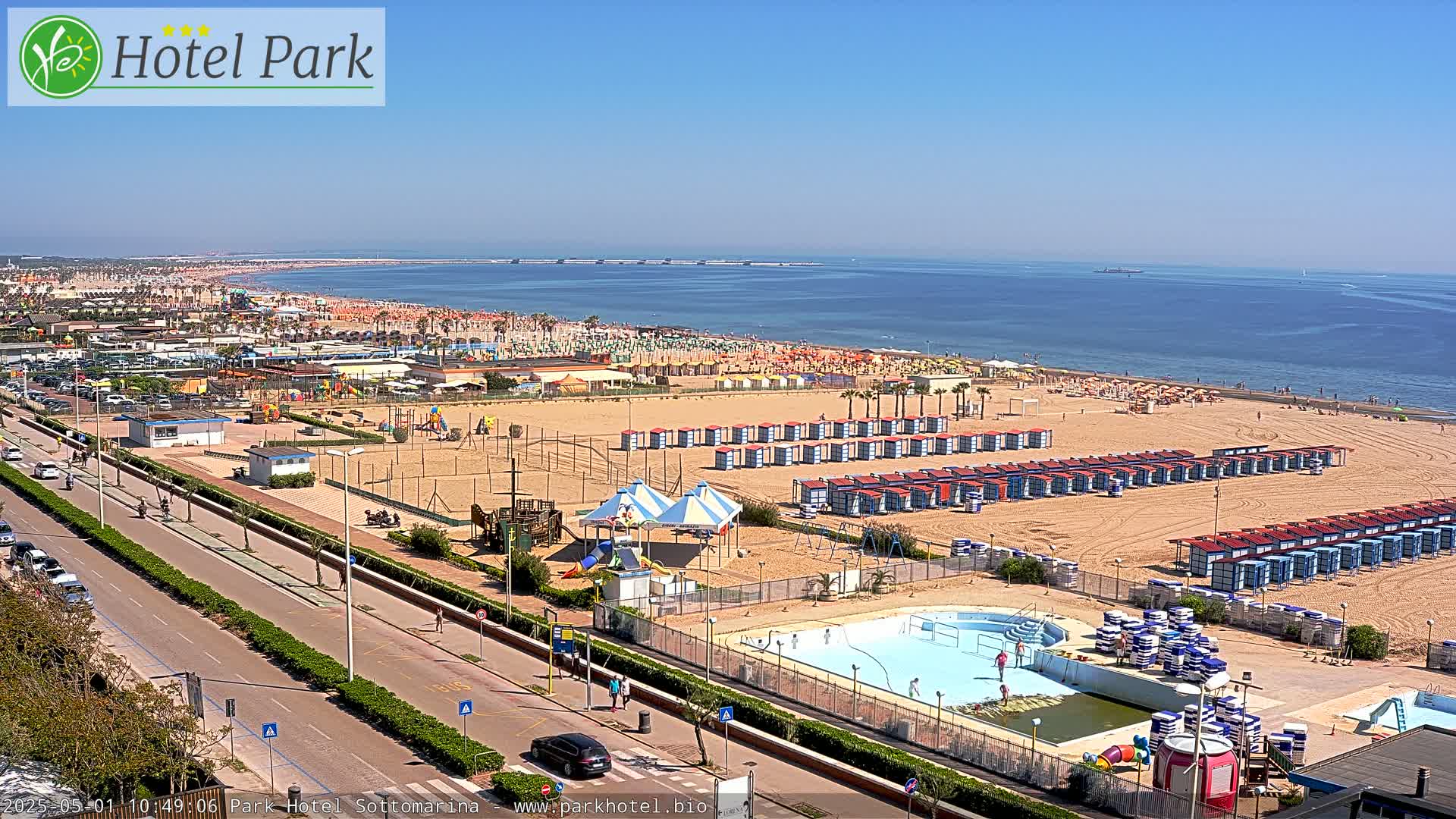 A sandy beach with numerous beach huts, a swimming pool, and playground equipment is visible, overlooking a calm ocean under a clear blue sky.