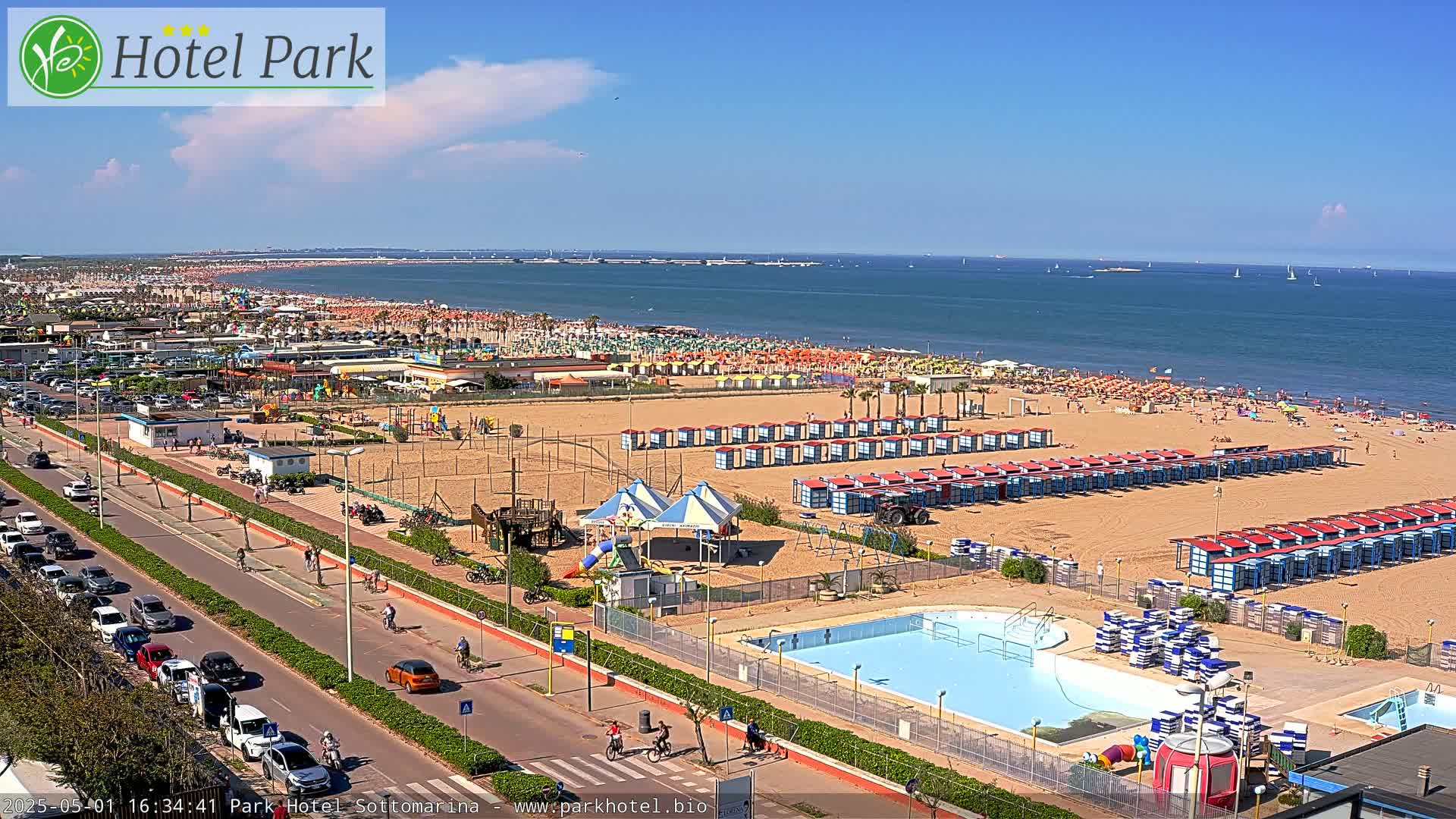 A sandy beach crowded with people and beach umbrellas, next to a road with cars and a swimming pool, on a sunny day.