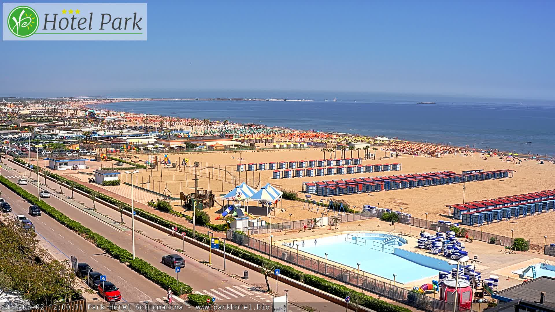 A sunny day at a beach with a swimming pool, beach cabanas, playground equipment, and a road with cars.