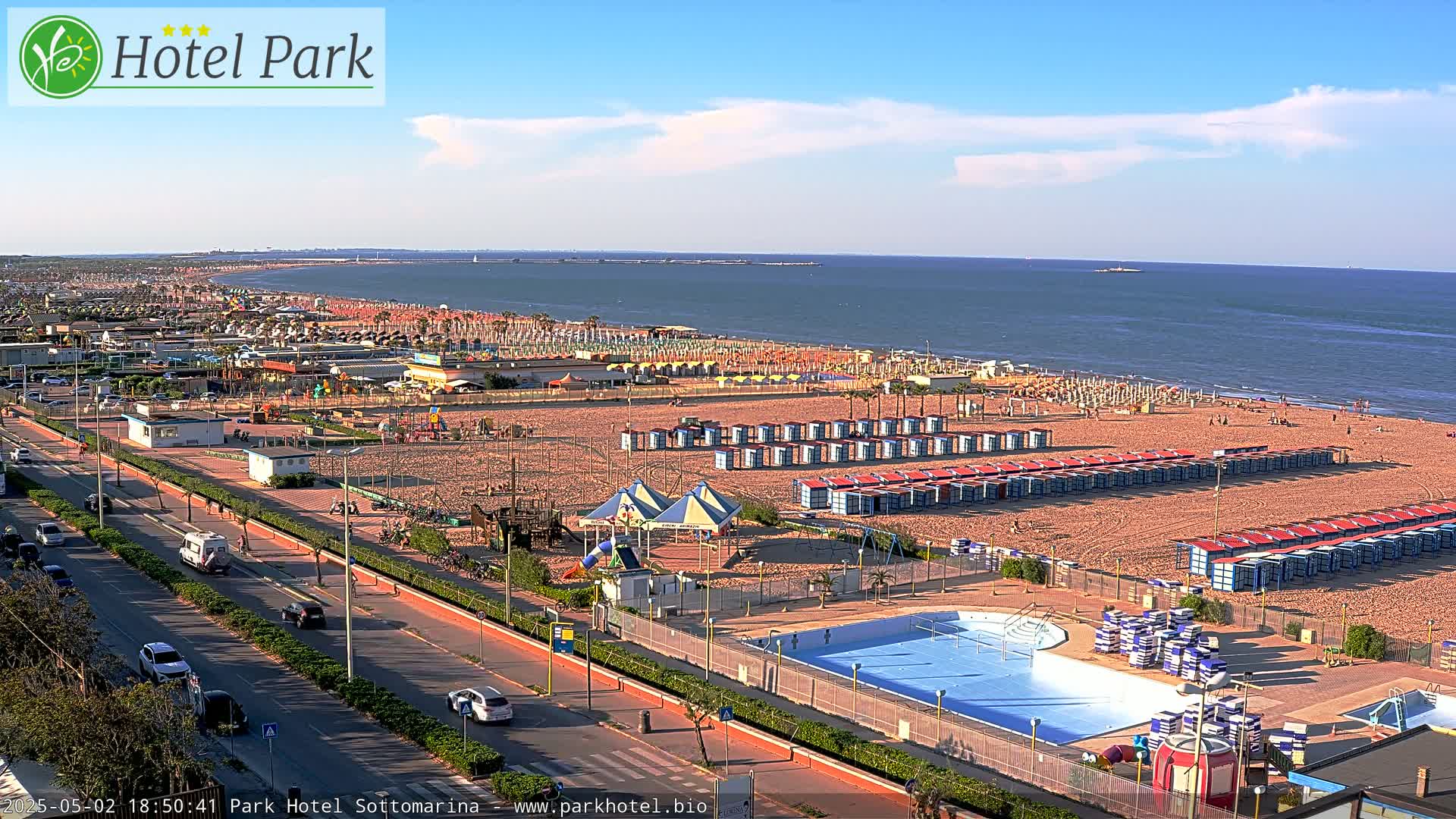 A sunny day overlooking a sandy beach with numerous beach umbrellas, changing rooms, a swimming pool, and a playground, all situated next to a road with cars.