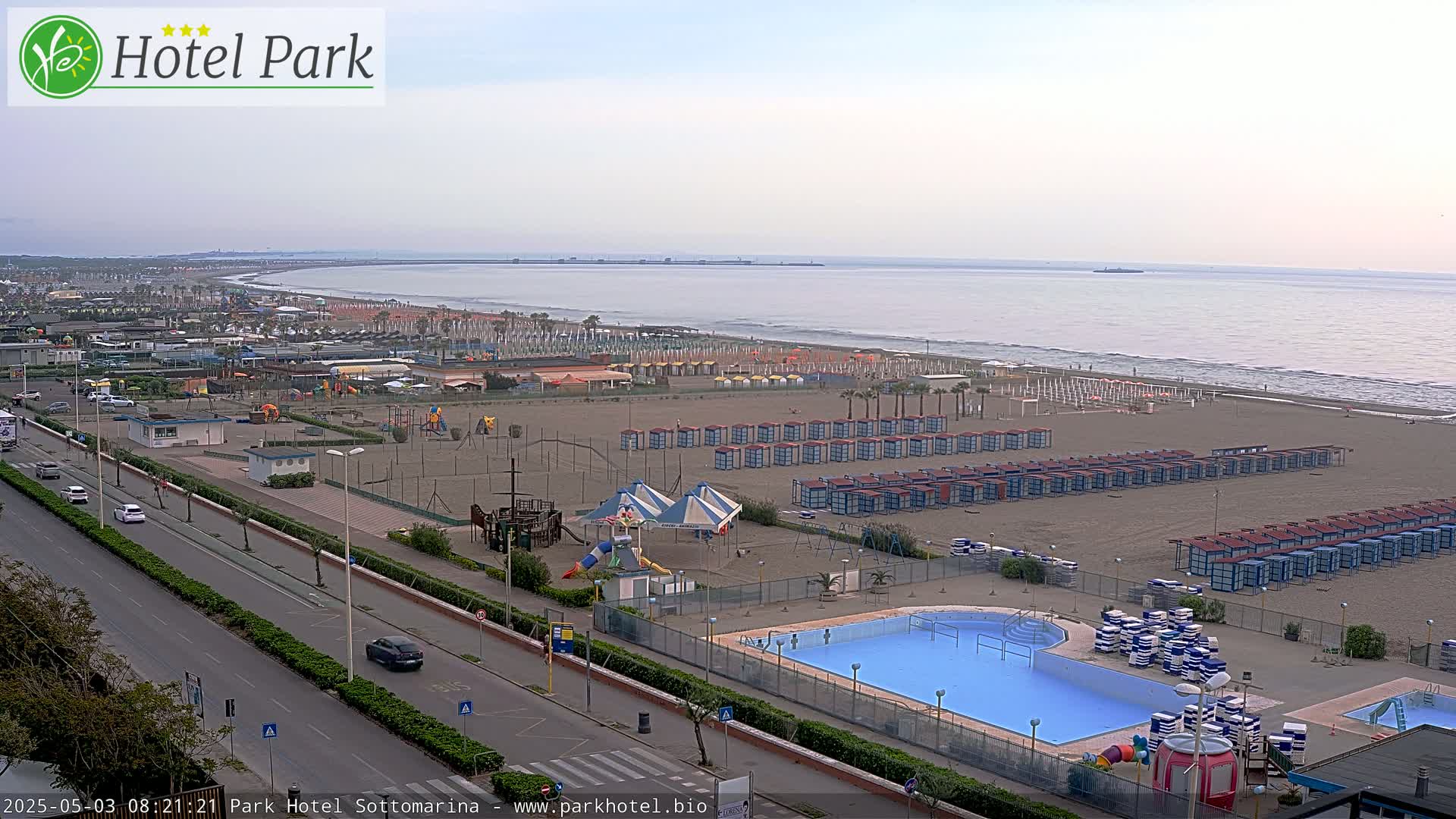 An outdoor view shows a beach with numerous beach huts, a swimming pool, playground equipment, and a roadway, all under a hazy, overcast sky.