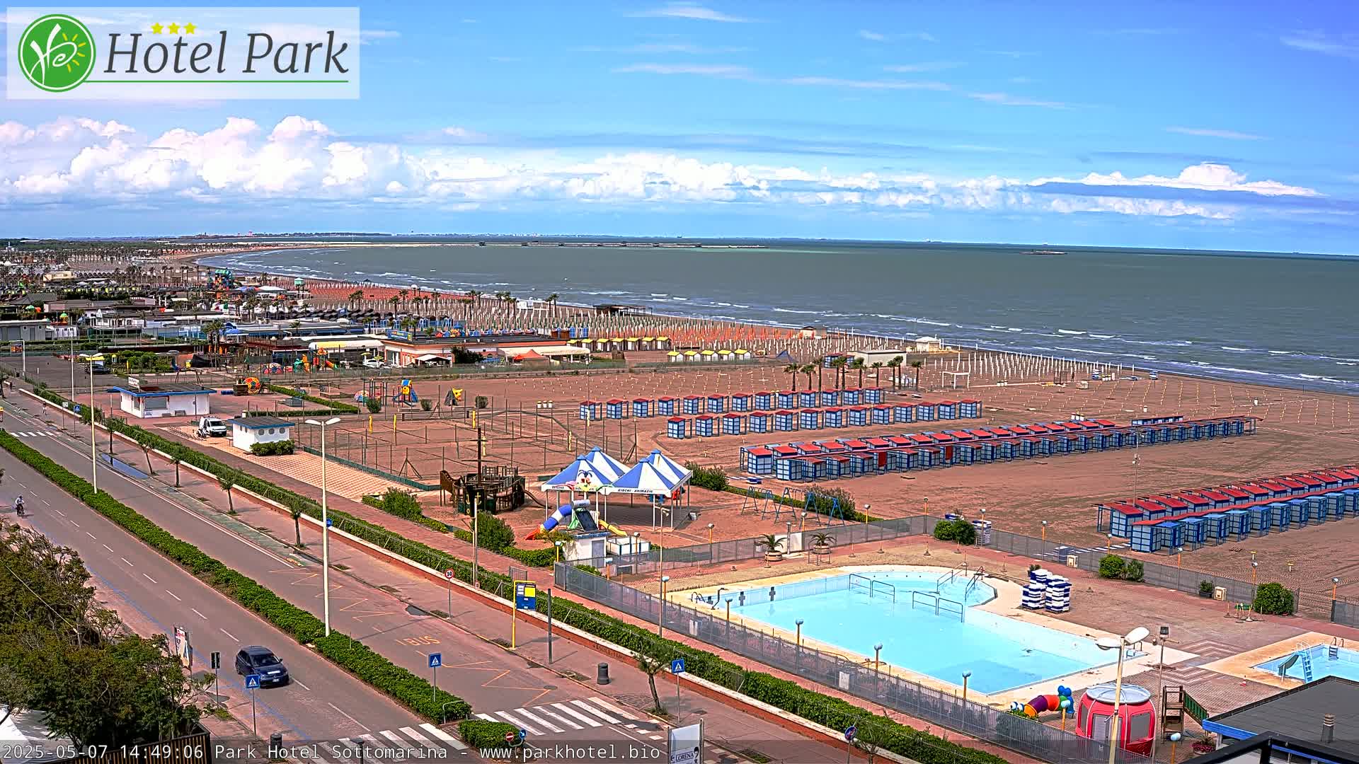 A sandy beach with rows of beach cabanas, a swimming pool, and playground equipment is visible, under a partly cloudy sky, next to a calm ocean.