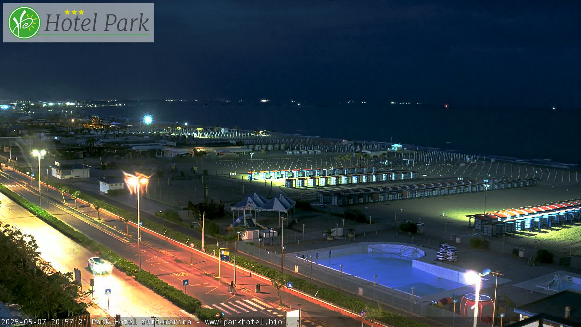 A nighttime beach scene shows rows of beach cabanas and a swimming pool next to a road, all illuminated under a dark sky.