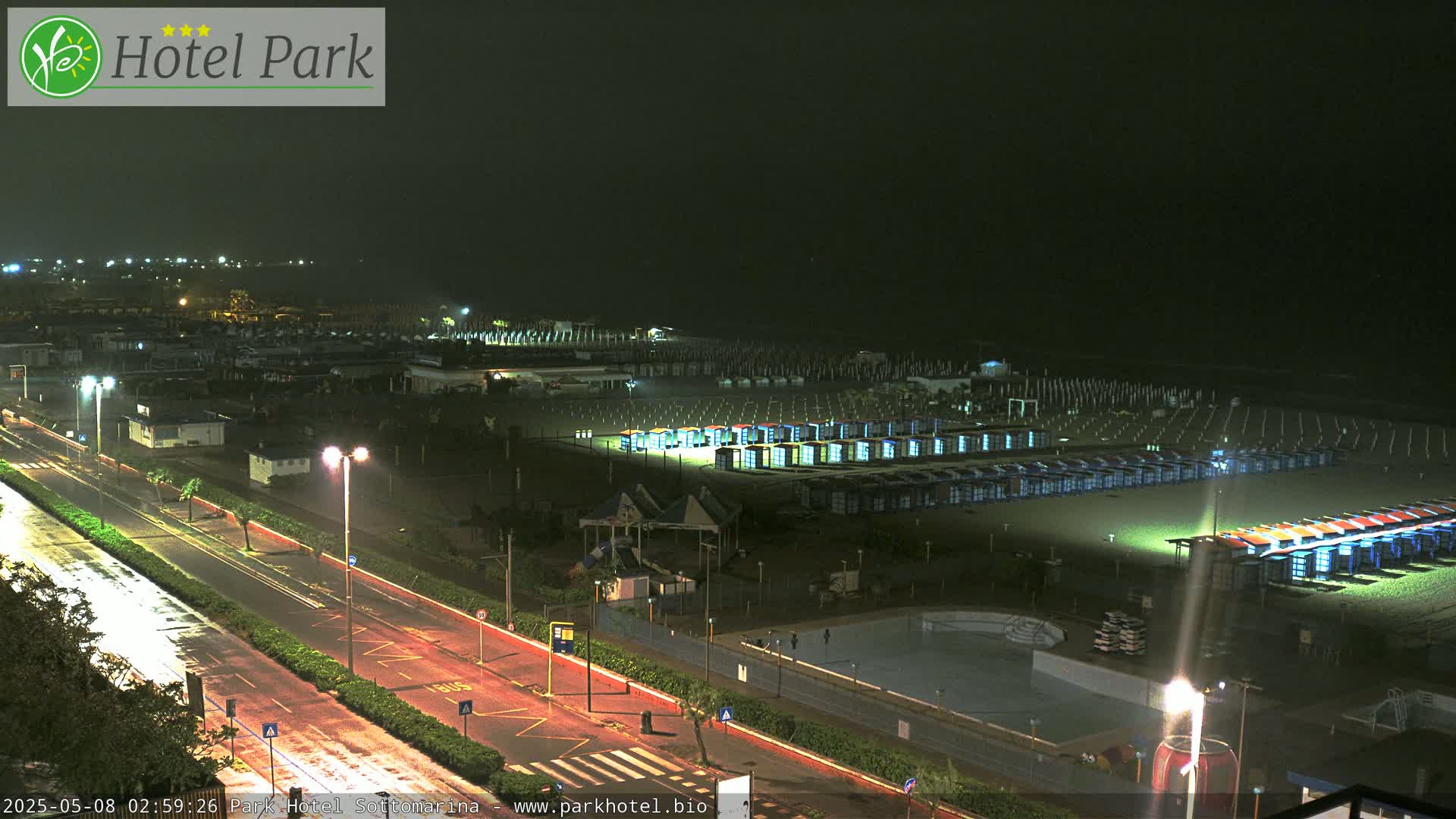A rain-slicked road runs alongside a beach at night, lined with rows of beach huts under a dark sky.