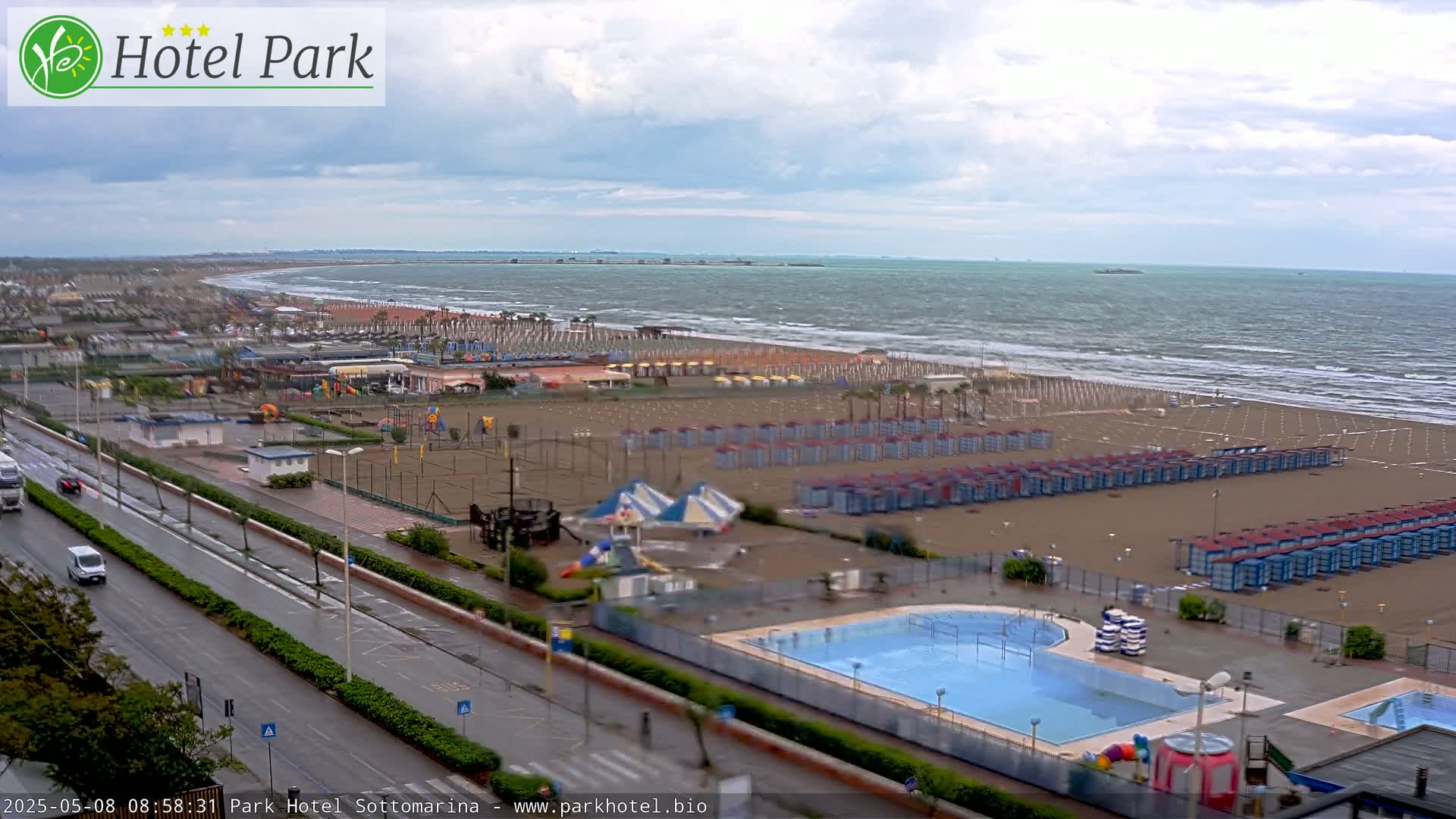 A beach with numerous beach huts and a swimming pool is shown under an overcast sky, with a road and buildings visible in the foreground.