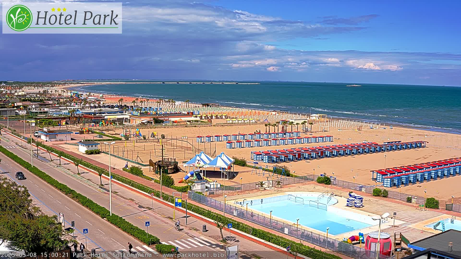 A sandy beach with rows of beach huts and a swimming pool is adjacent to a road with some cars and pedestrians, all under a partly cloudy sky.