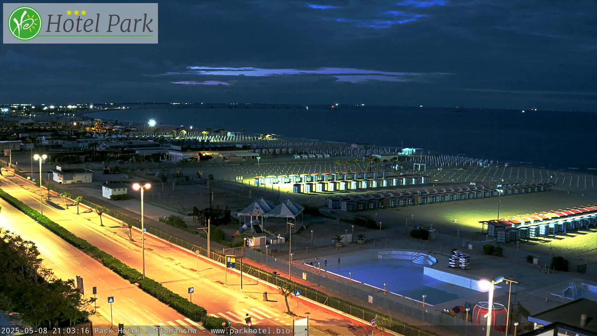 A nighttime view of a beach with rows of beach cabanas, a pool, and a road, all lit by streetlights under a dark, cloudy sky.