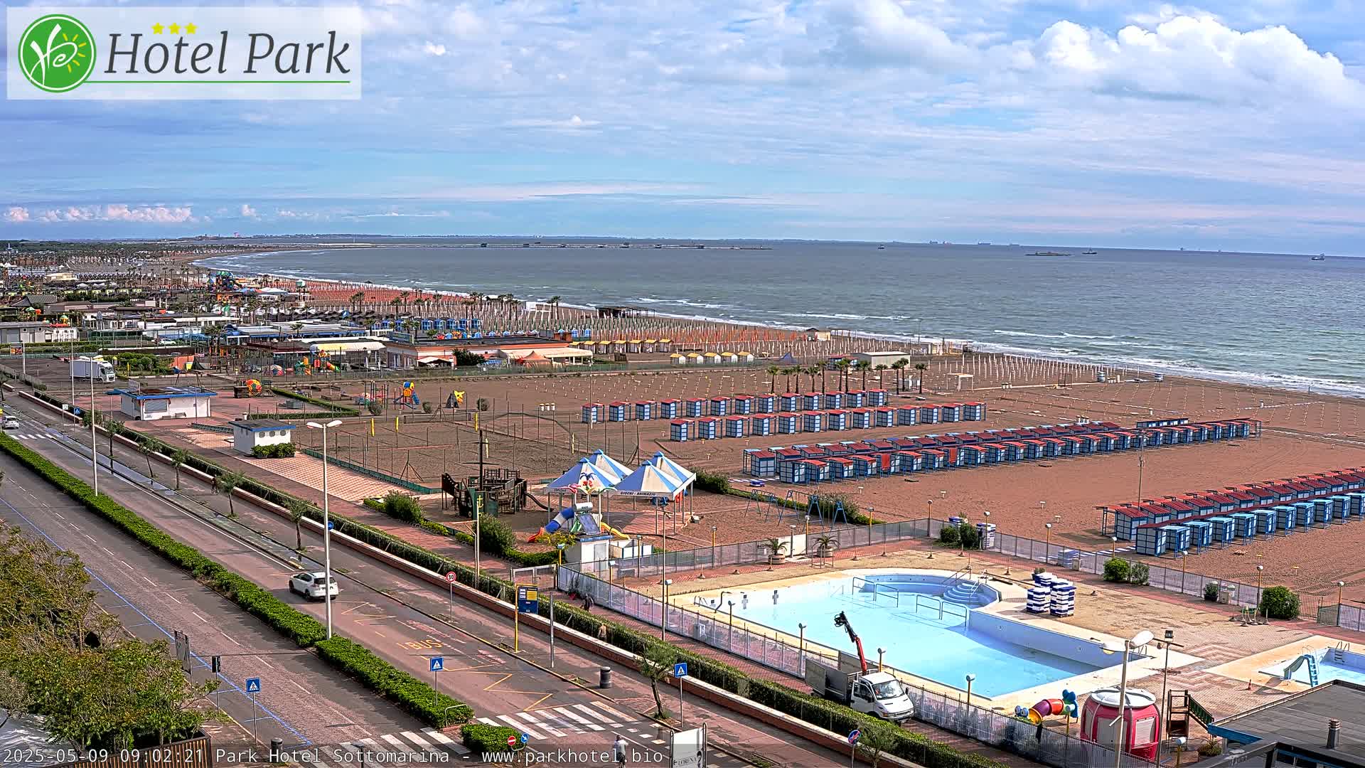 A sandy beach with rows of beach huts, a swimming pool, a playground, and a road, all under a partly cloudy sky and next to a calm sea.