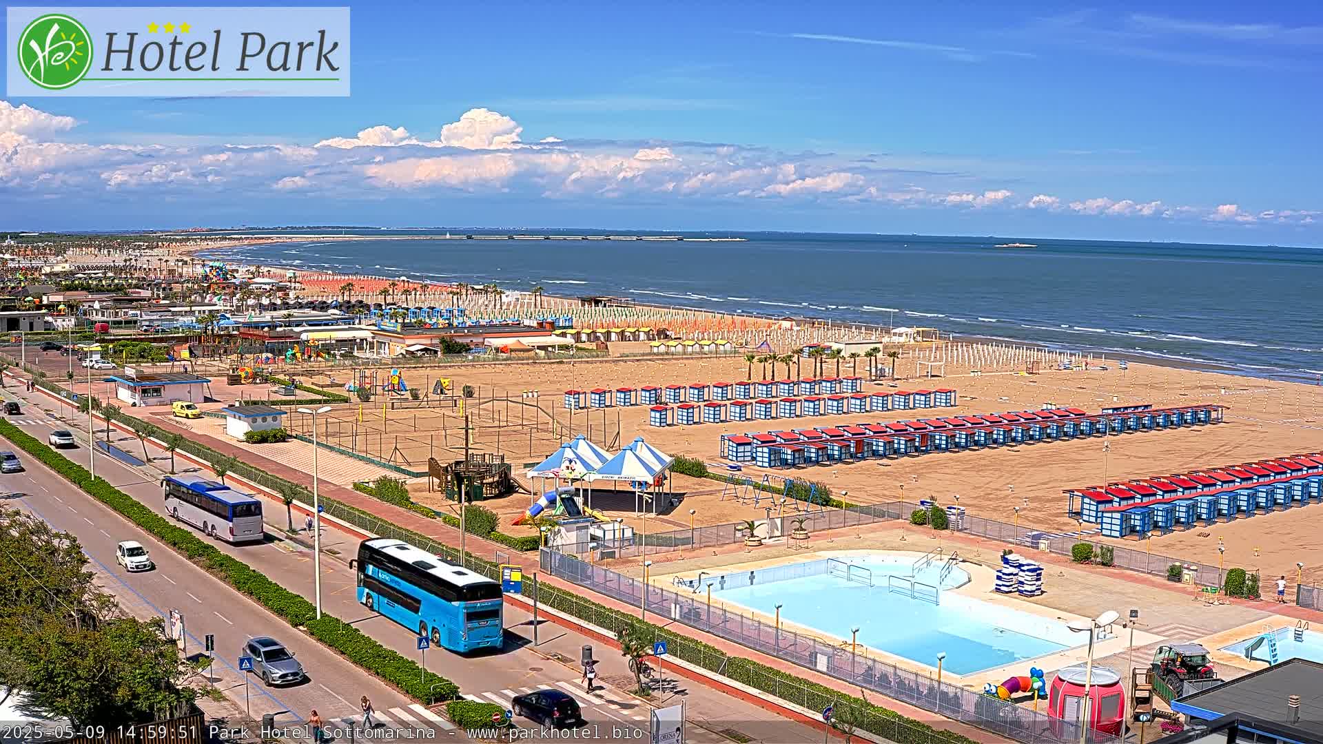 A sandy beach with rows of beach huts, a swimming pool, playground equipment, and a road with buses is visible on a partly cloudy day.