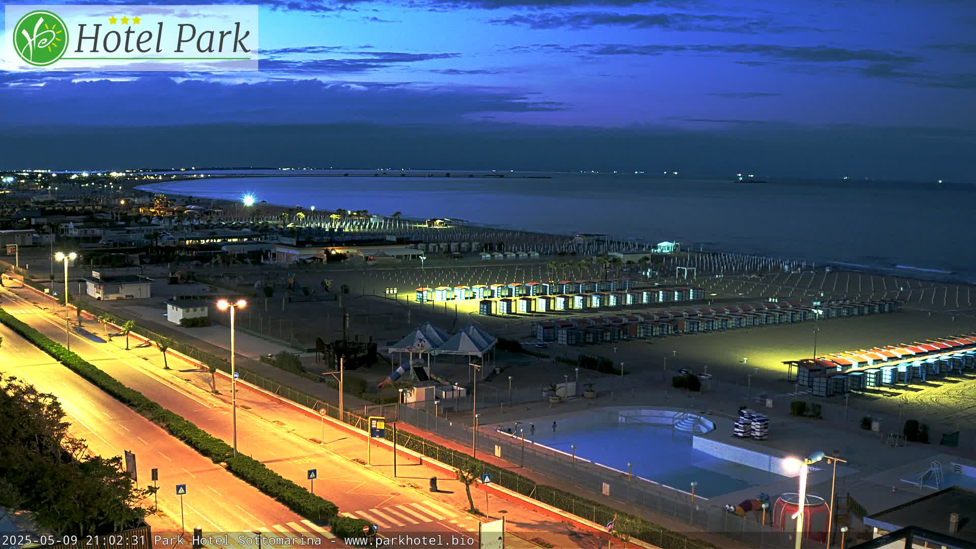 A nighttime view of a beach with rows of beach huts, a swimming pool, and a road illuminated by streetlights, under a dark blue twilight sky.