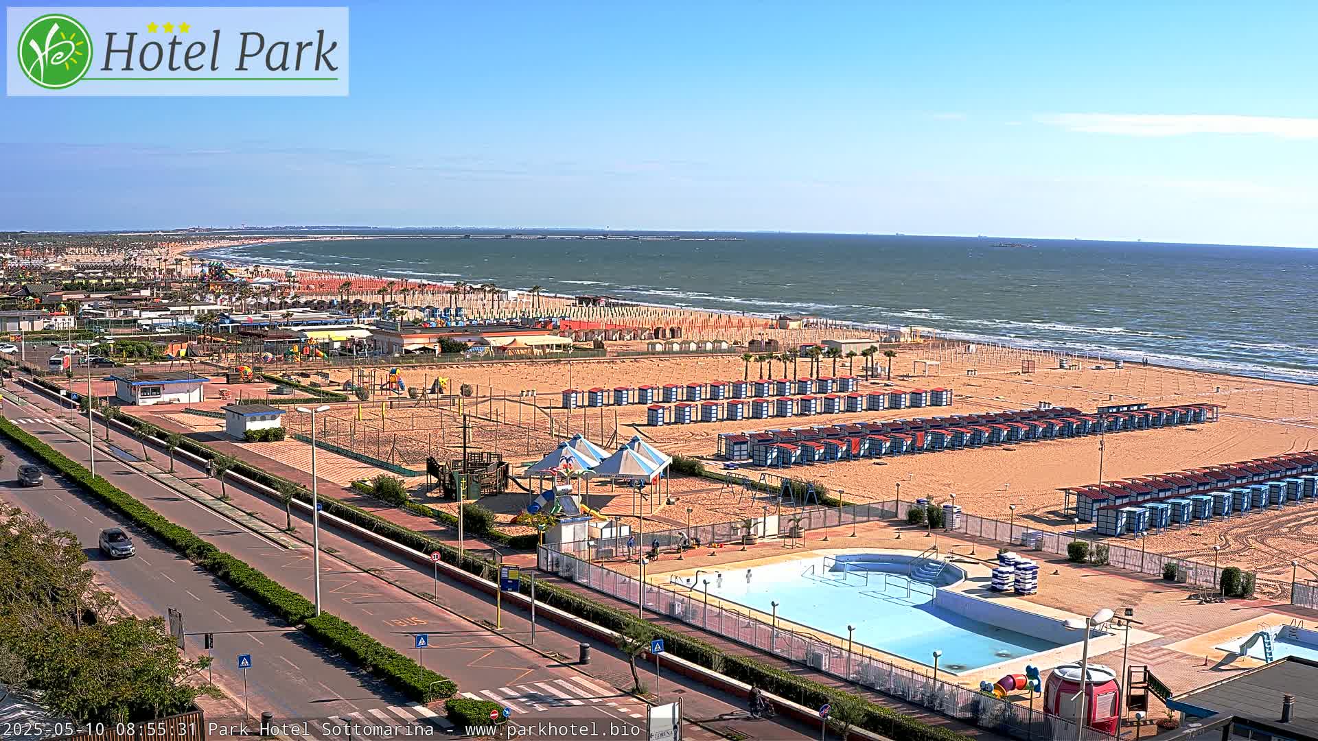 A sunny beach scene shows a sandy shoreline with rows of beach umbrellas and chairs, a swimming pool, and a roadway, all overlooking a calm ocean.