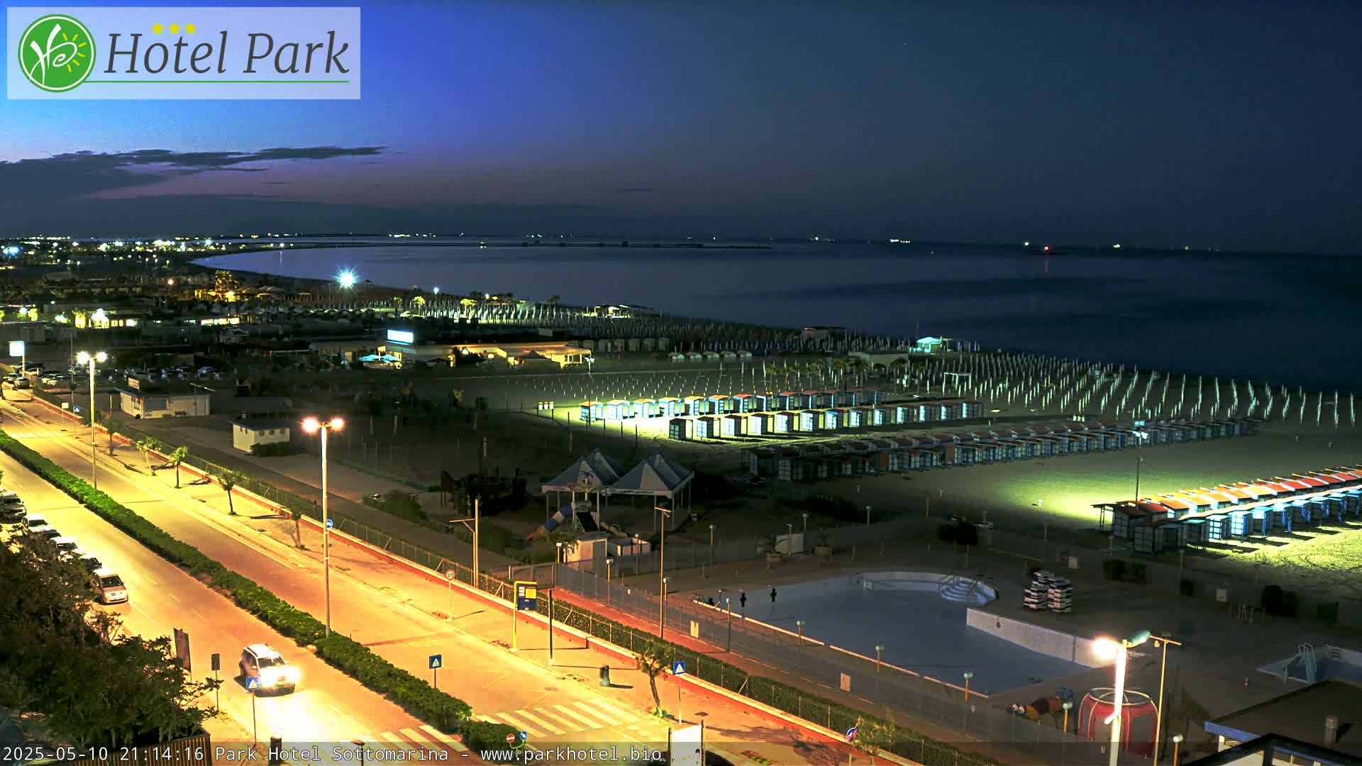A nighttime view of a beach with rows of beach huts, a road with cars, and a swimming pool, under a dark, clear sky.