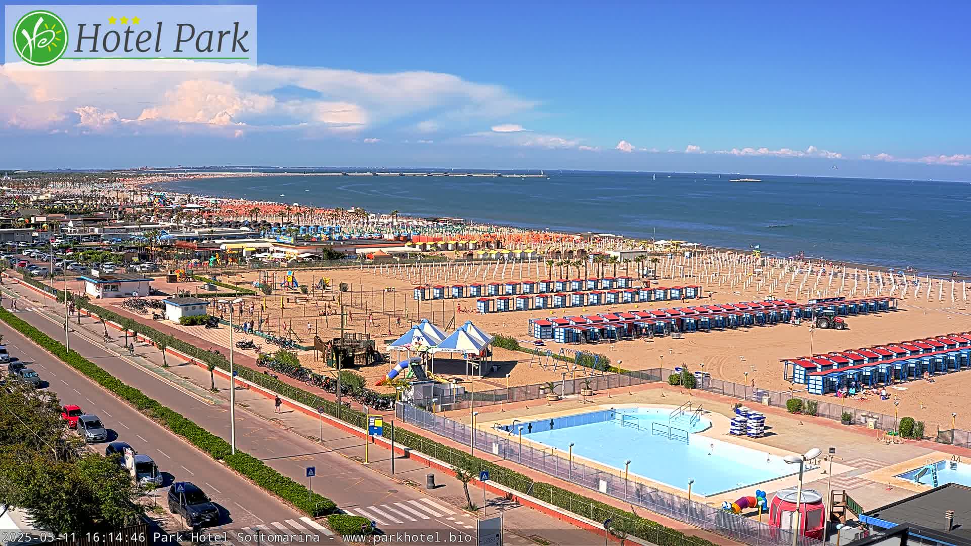 A sandy beach with numerous beach umbrellas and cabanas, a swimming pool, and a playground, all under a sunny sky with a few clouds.