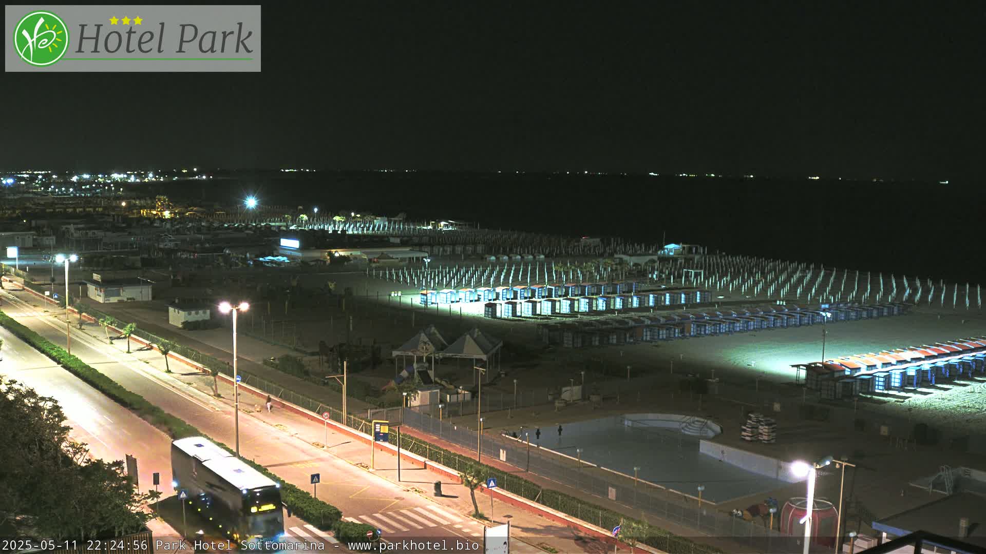 A nighttime view of a beach with rows of beach huts illuminated, a road with a bus, and a swimming pool visible in the foreground.