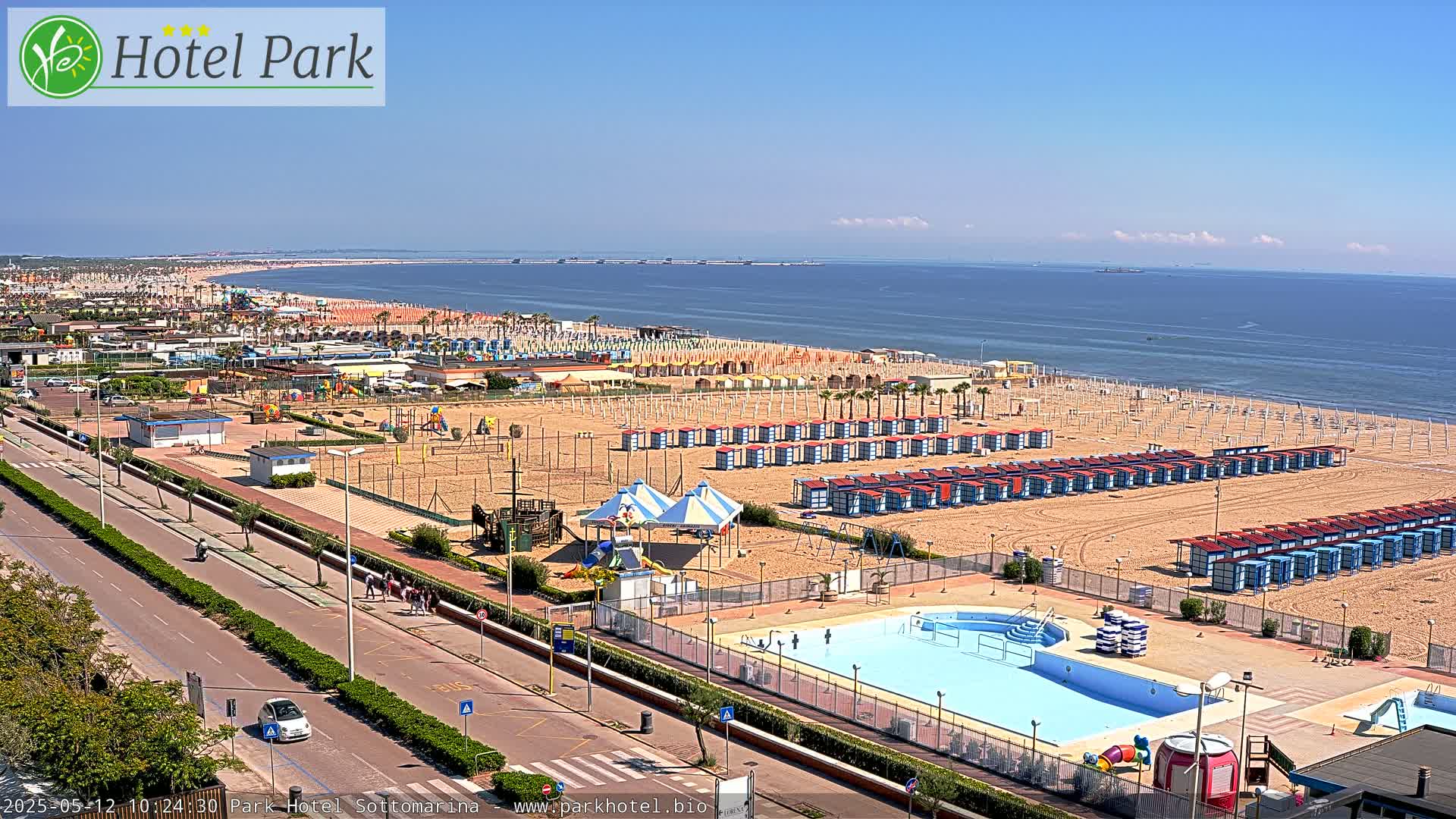 A beach scene on a sunny day shows a sandy beach with numerous beach huts, a swimming pool, playground equipment, and a road leading to the ocean.