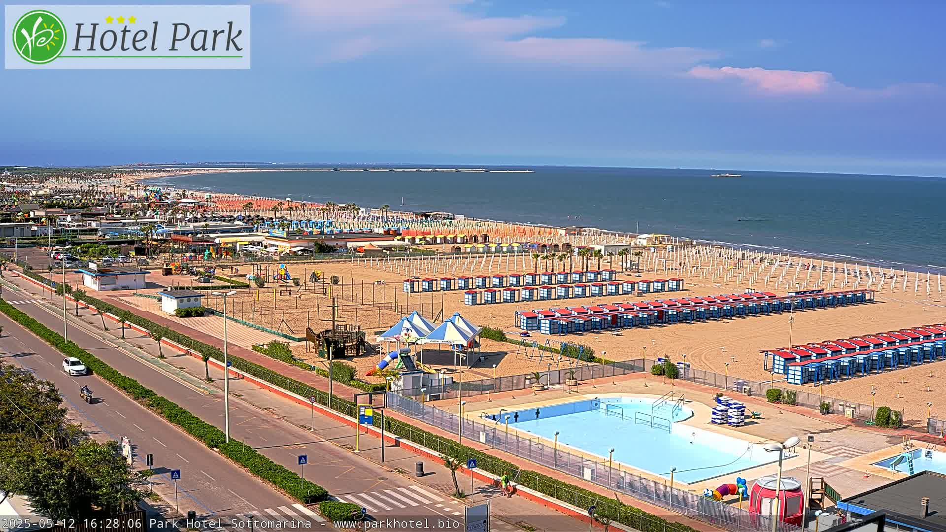 A sunny day at a beach resort shows a sandy beach lined with rows of beach umbrellas and cabanas, a swimming pool, playground equipment, and a road running alongside.