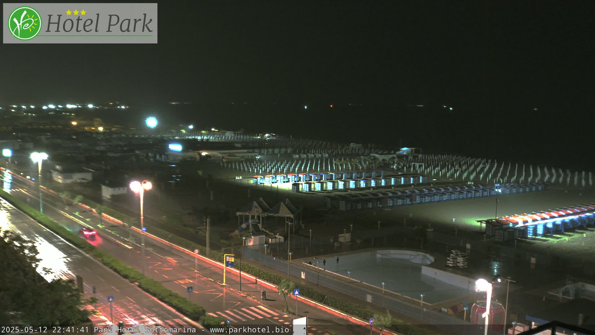 A rain-slicked road runs alongside a dark beach at night, lined with rows of beach huts under dim lighting.