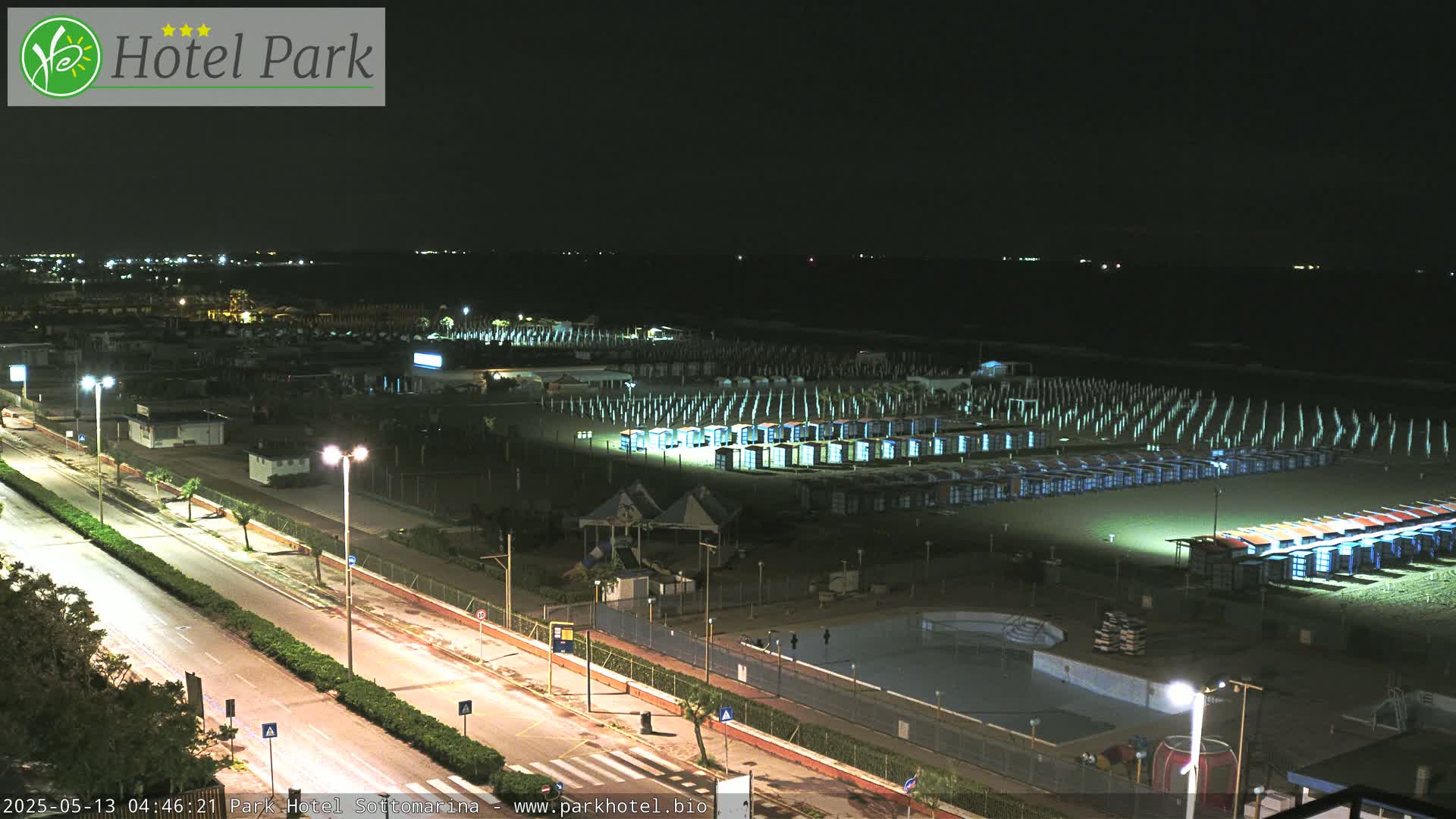 A nighttime aerial view shows a beach with rows of beach cabanas illuminated, a road, and a pool area.