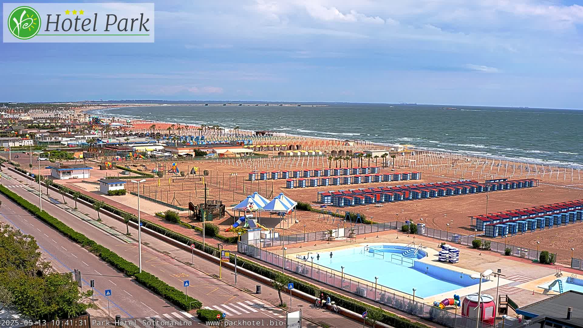 A beach with numerous beach huts and a swimming pool is seen from an elevated viewpoint, under a partly cloudy sky.