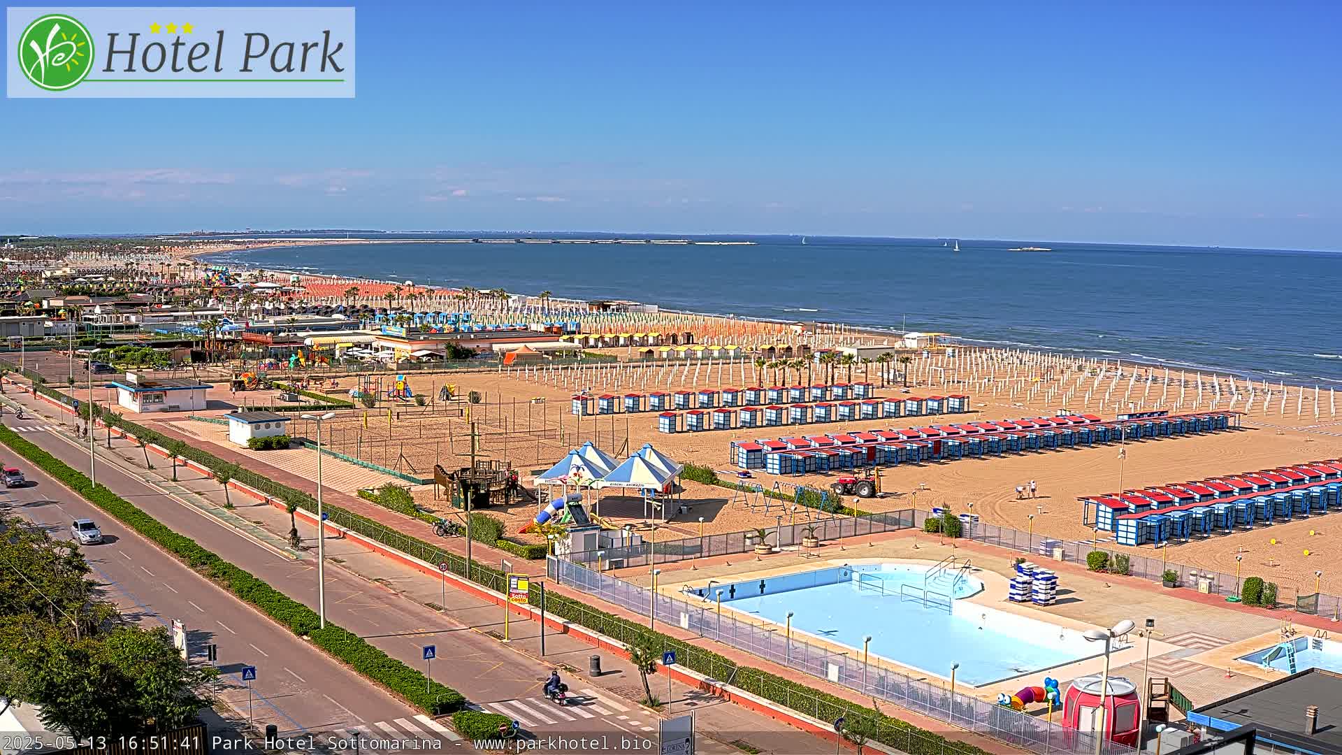 A sandy beach with rows of beach umbrellas and changing rooms, a swimming pool, playground equipment, and a road bordering the ocean on a sunny day.