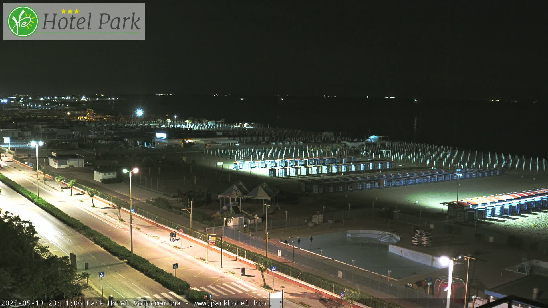 A nighttime view of a beach with rows of beach huts illuminated along a roadway.