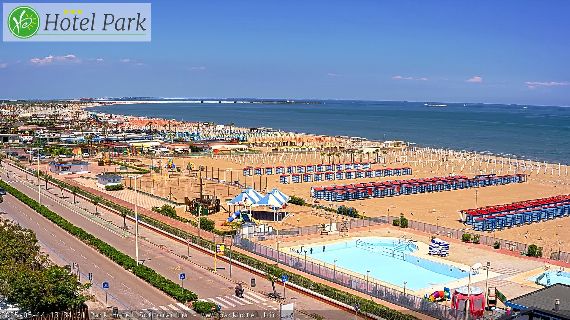 A sunny day at a beach resort shows a sandy beach with rows of beach huts, a swimming pool, playground equipment, and a roadway leading to the ocean.
