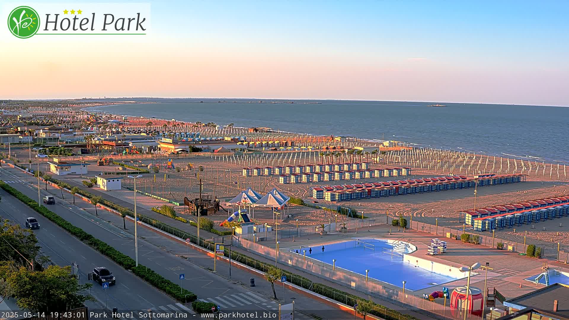 A sandy beach with rows of beach umbrellas and changing rooms, a swimming pool, playground equipment, and a road with cars, all under a clear, sunset sky.